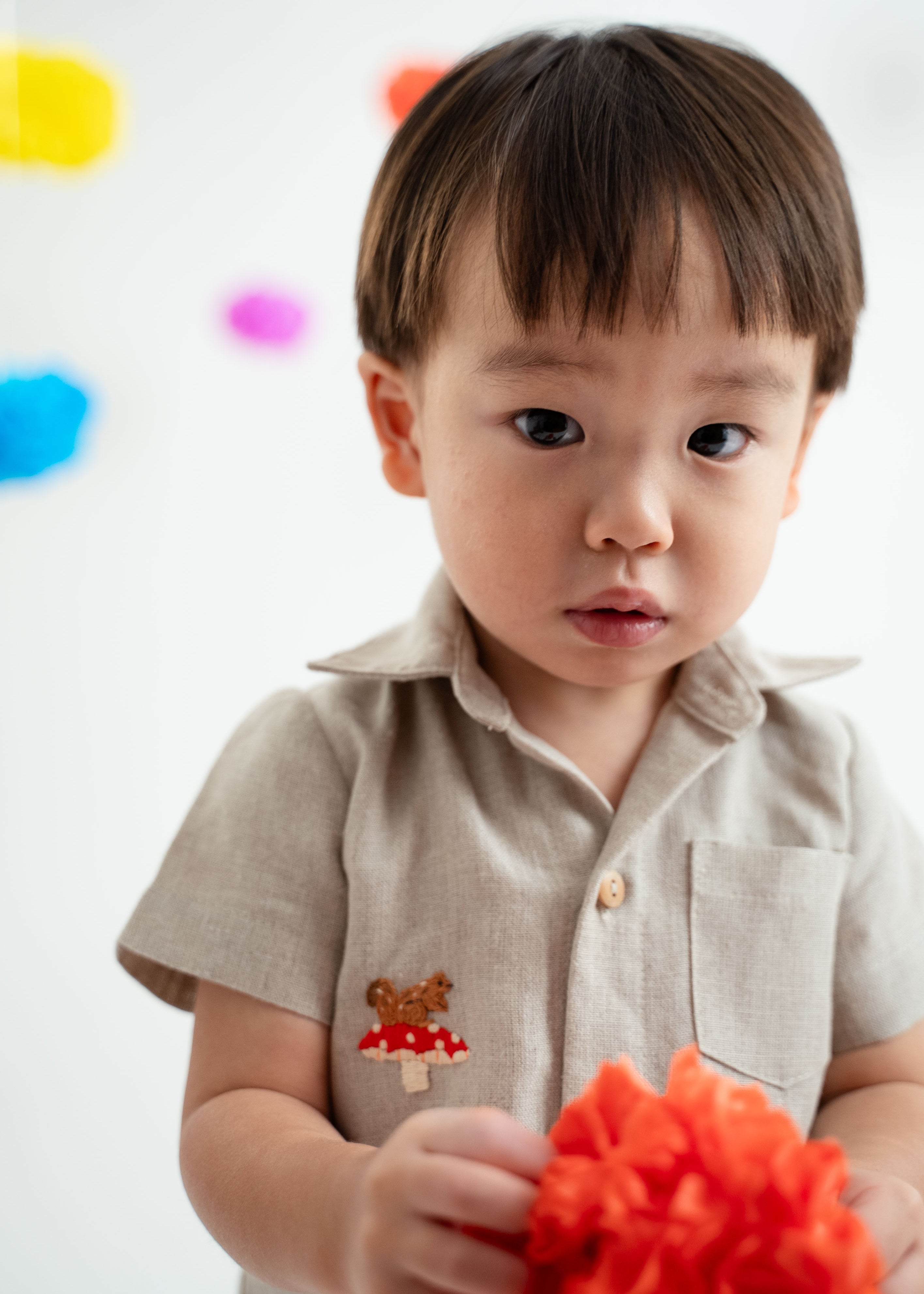 Close de menino vestindo macacão infantil unissex em linho cru com bordado de cogumelo e esquilo.
