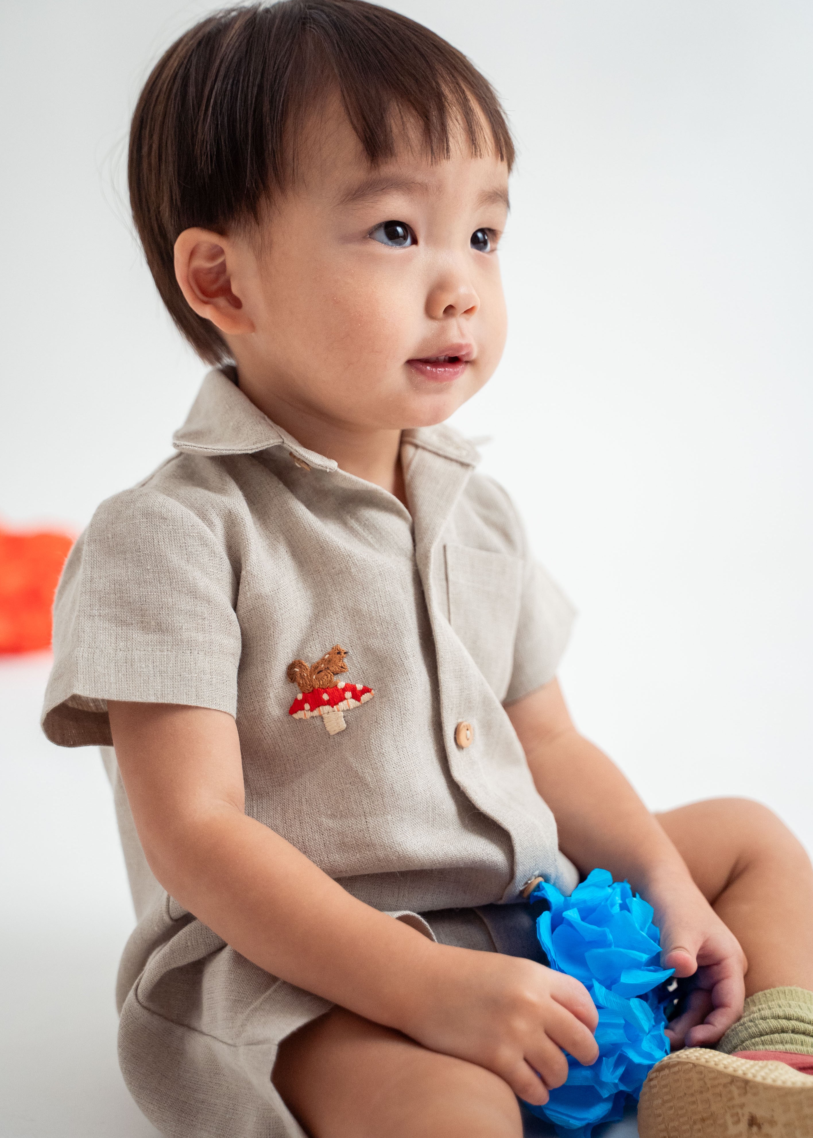 Menino sentado vestindo macacão infantil unissex em linho cru com bordado de cogumelo e esquilo.
