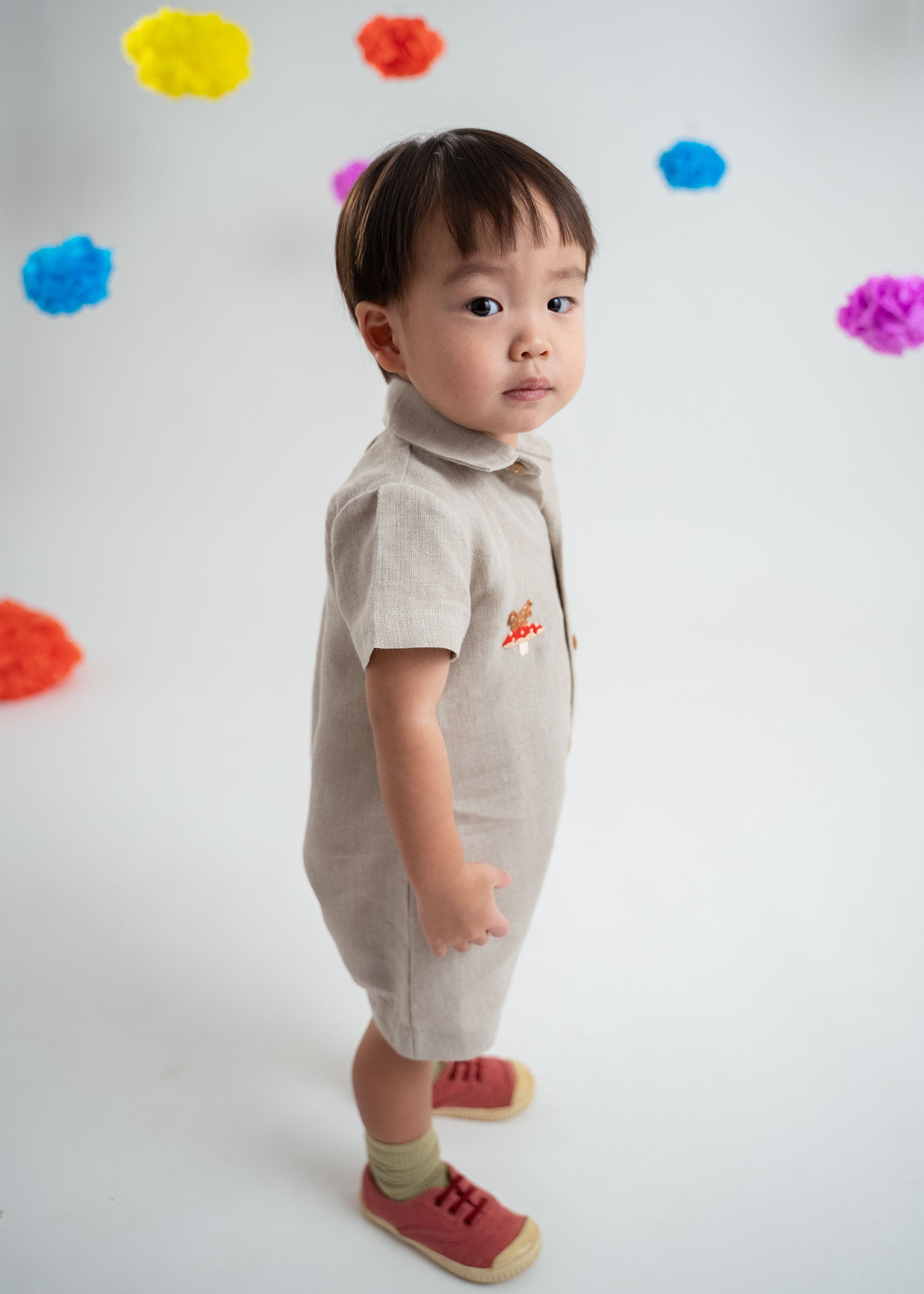 Menino usando macacão infantil unissex em linho cru com bordado de cogumelo e esquilo.