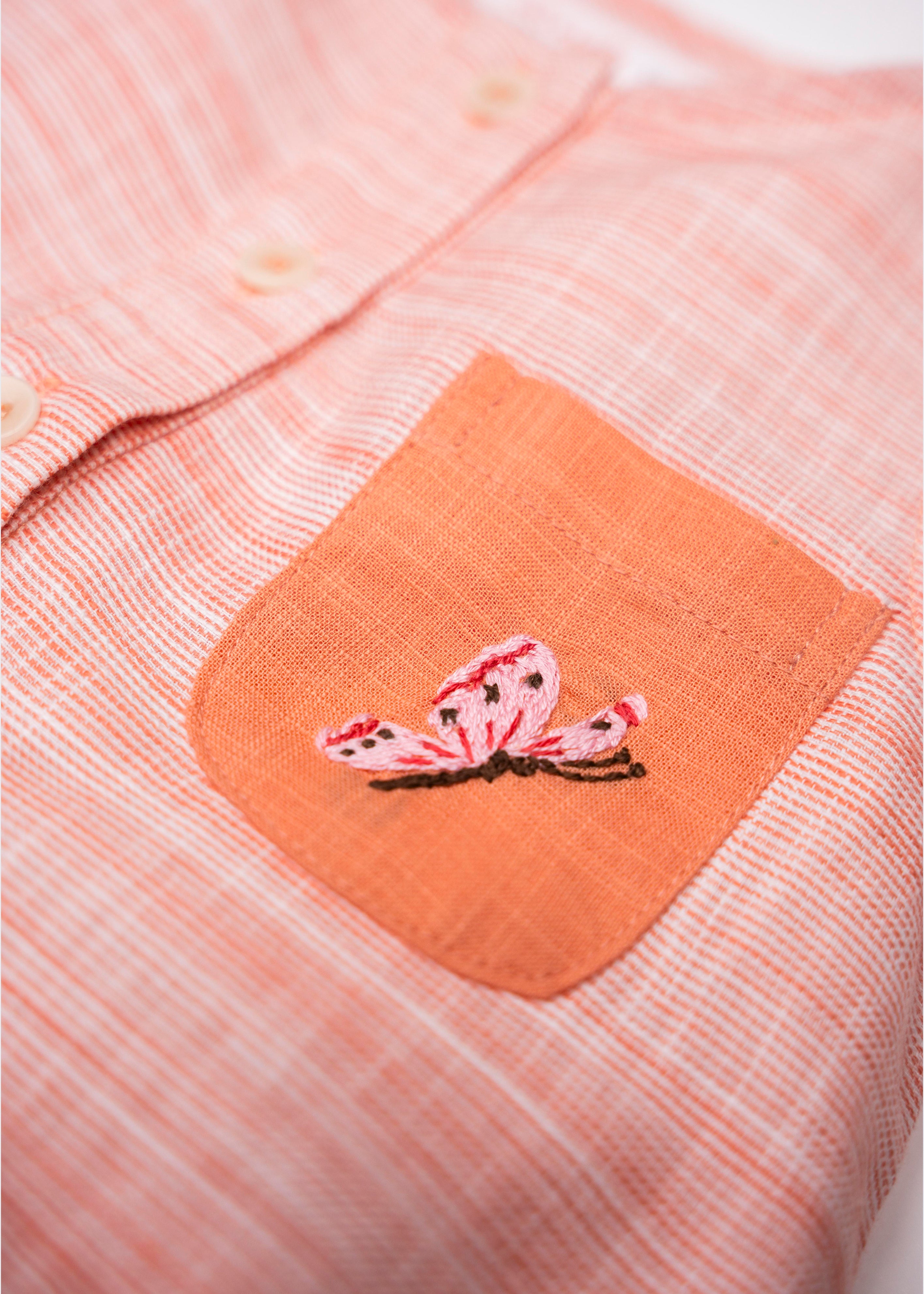 Detalhe da borboleta rosa bordada à mão no bolso laranja do Macaquinho Valen em algodão orgânico.