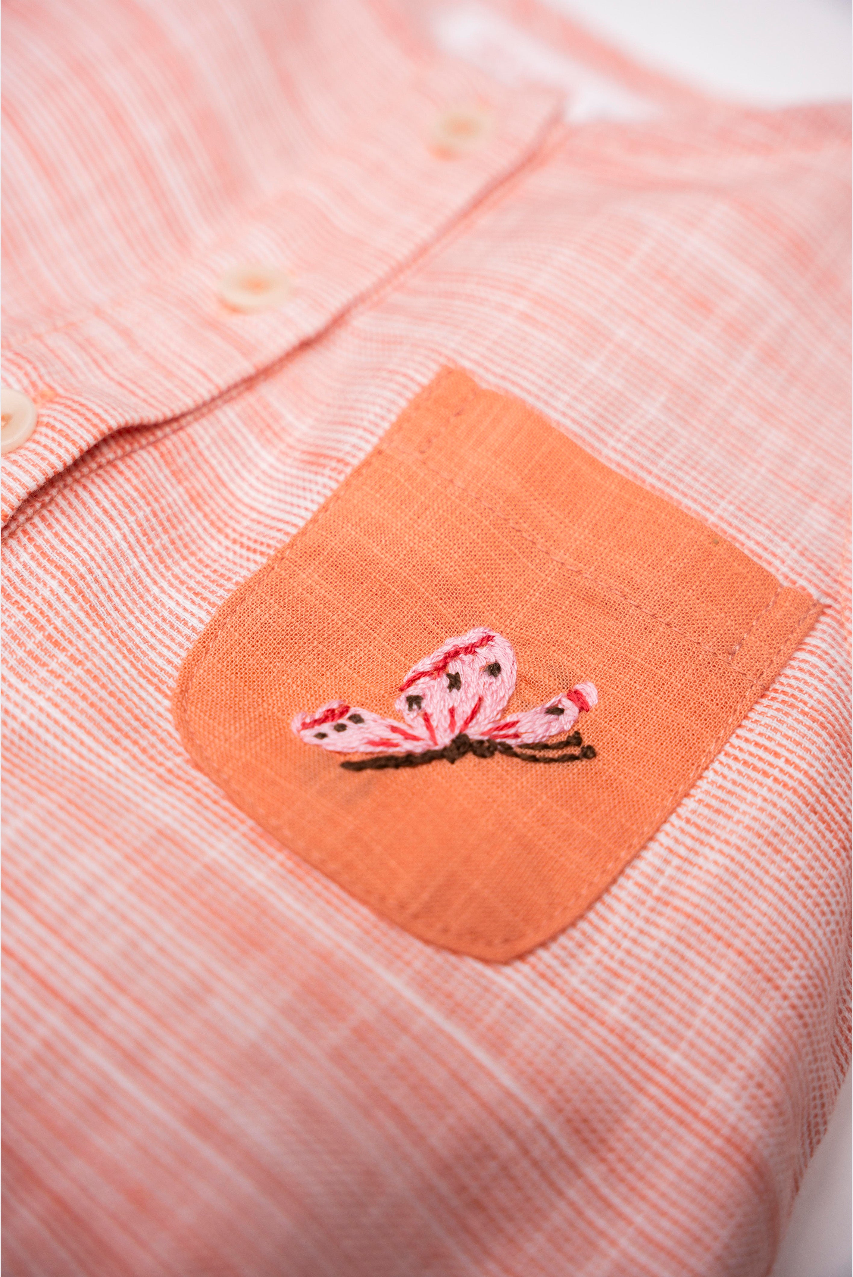 Detalhe da borboleta rosa bordada à mão no bolso laranja do Macaquinho Valen em algodão orgânico.