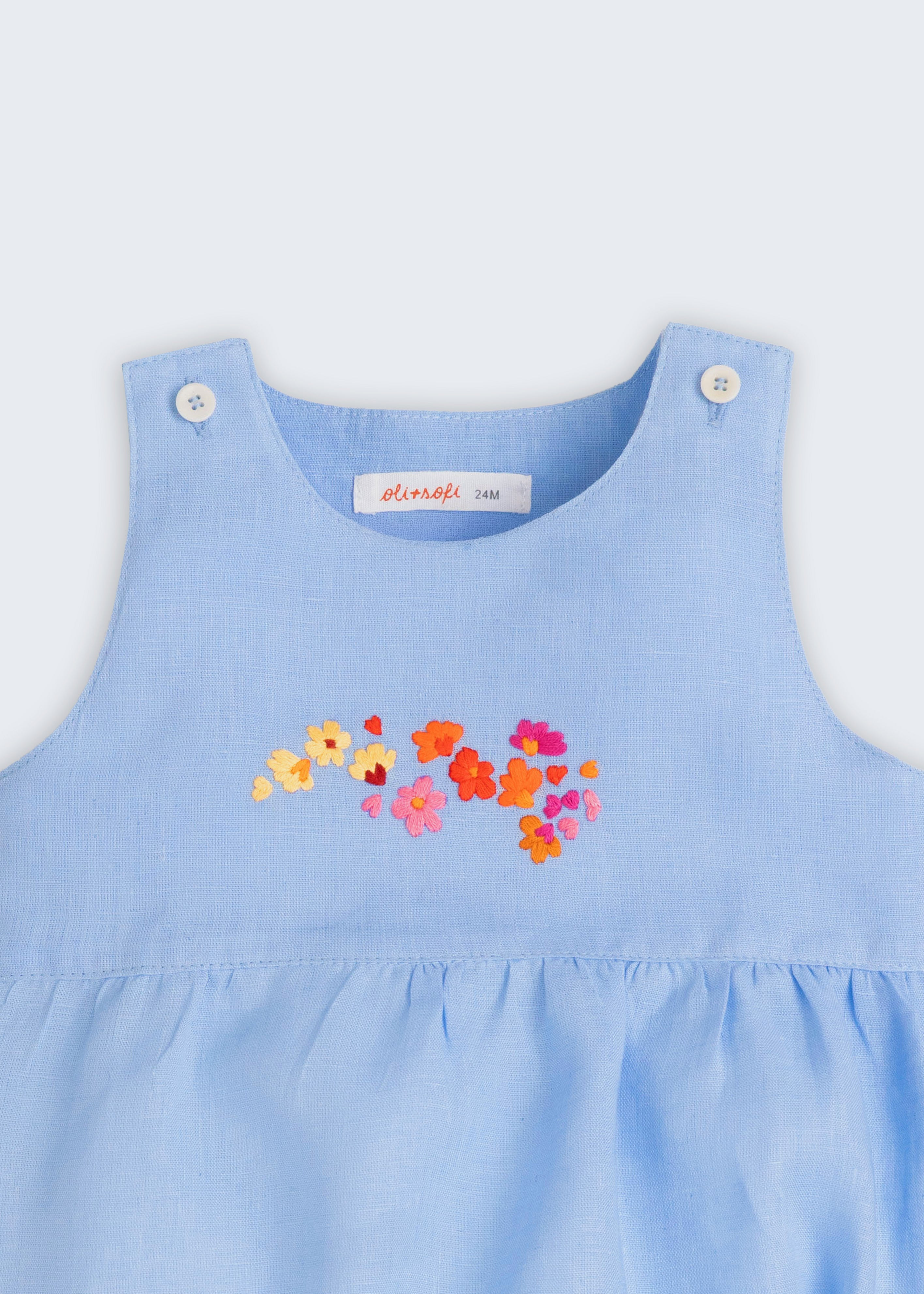 Macaquinho de bebê estilo romper unissex em linho azul com bordado de flores, visto de frente em detalhe.