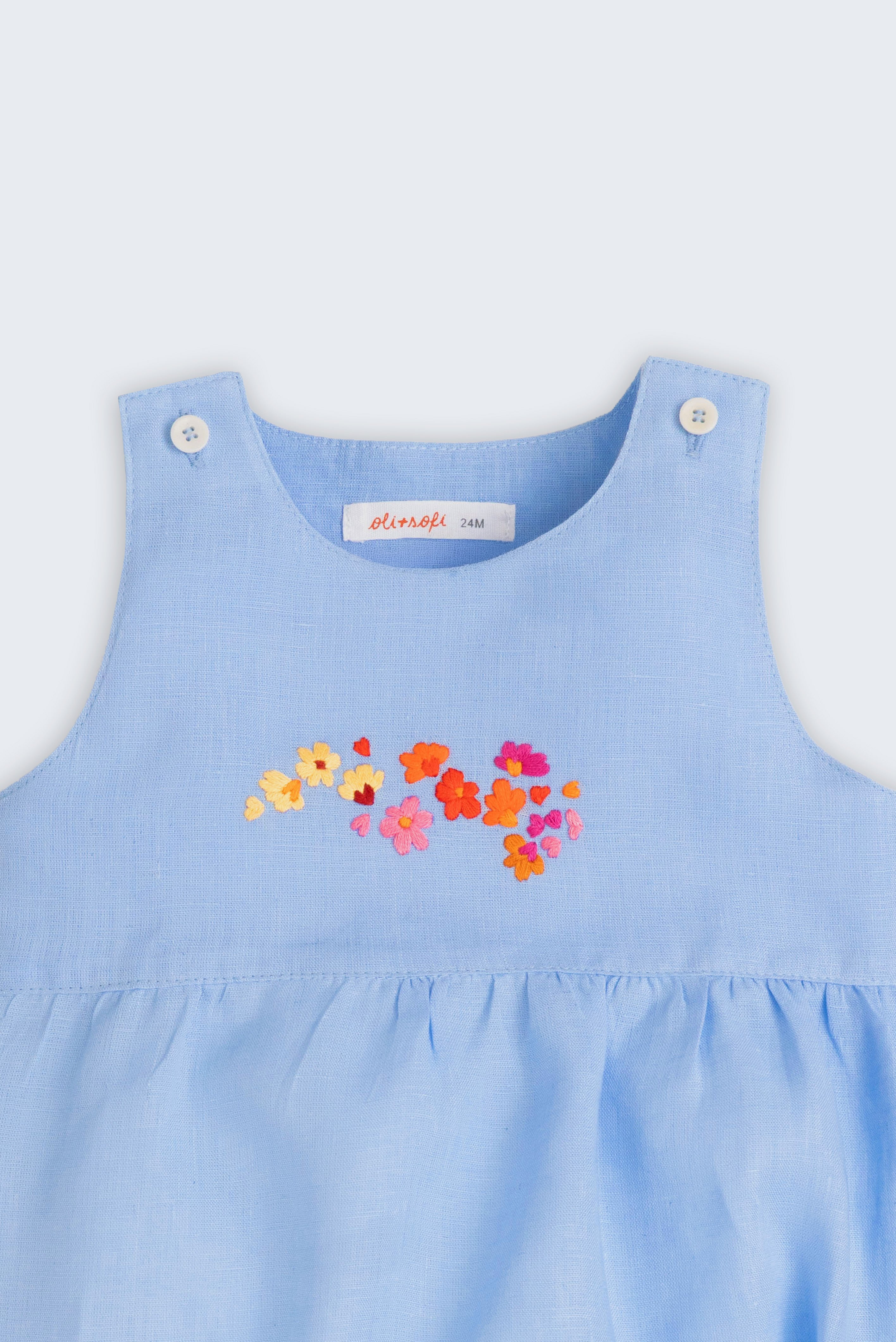 Macaquinho de bebê estilo romper unissex em linho azul com bordado de flores, visto de frente em detalhe.