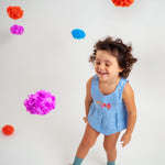 Menina sorrindo usando macaquinho estilo romper de bebê em linho azul-celeste com bordado de flores.