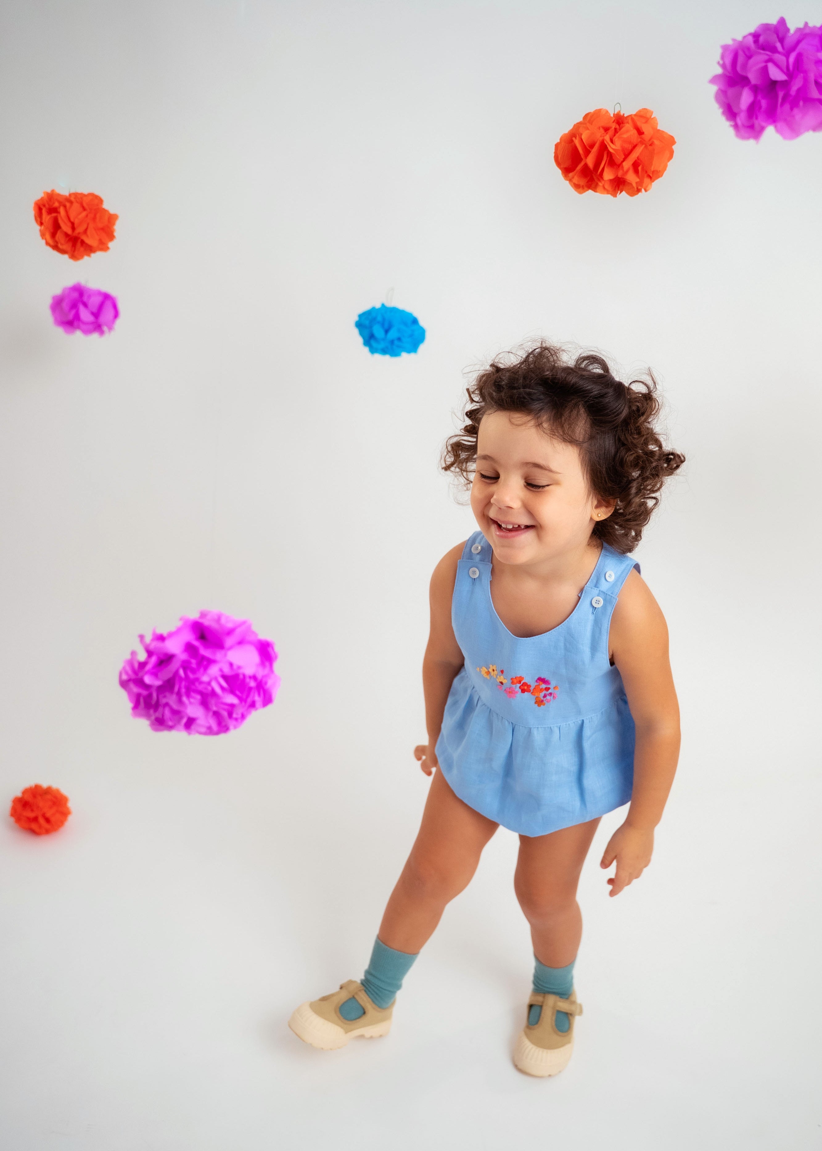 Menina sorrindo usando macaquinho estilo romper de bebê em linho azul-celeste com bordado de flores.
