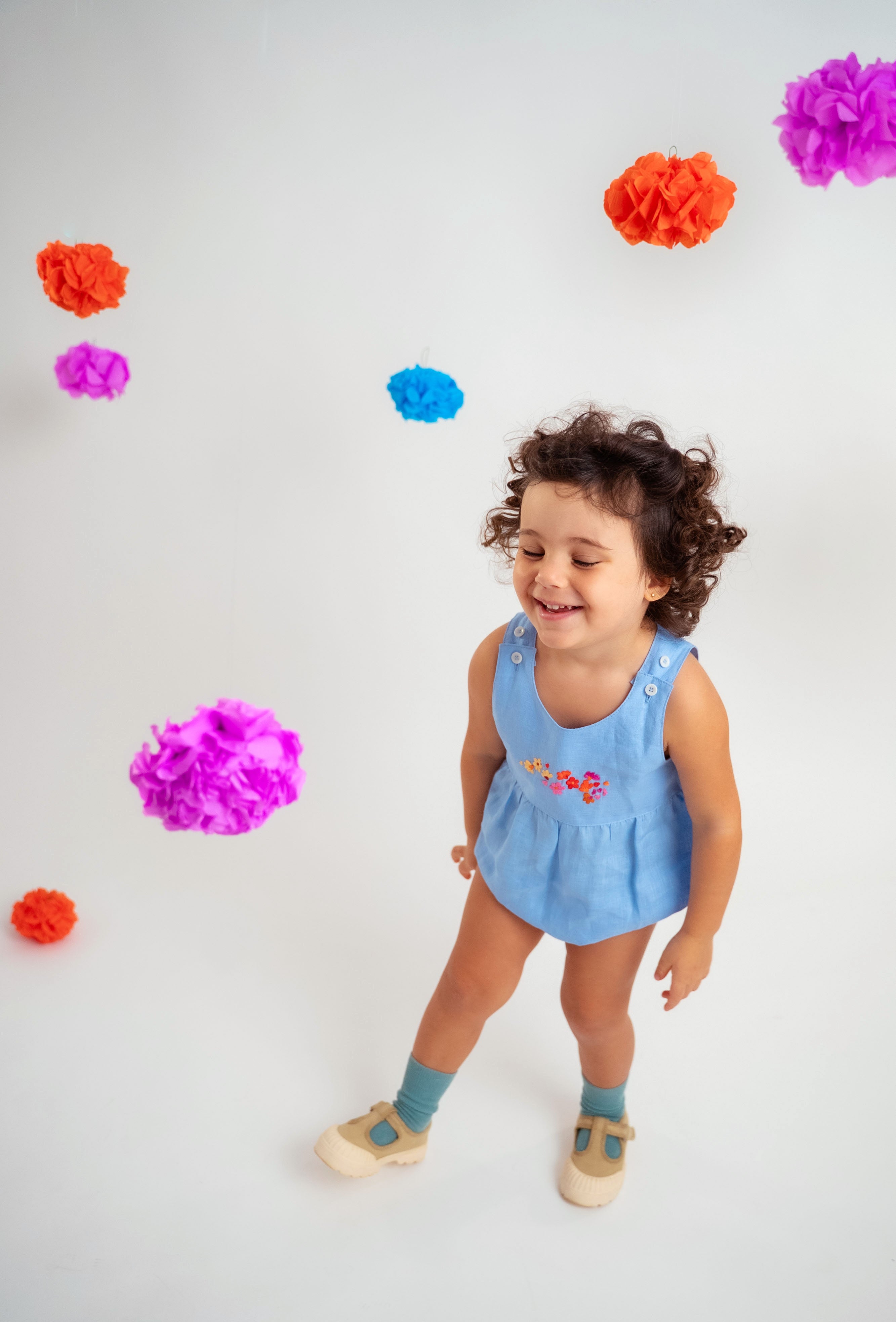 Menina sorrindo usando macaquinho estilo romper de bebê em linho azul-celeste com bordado de flores.