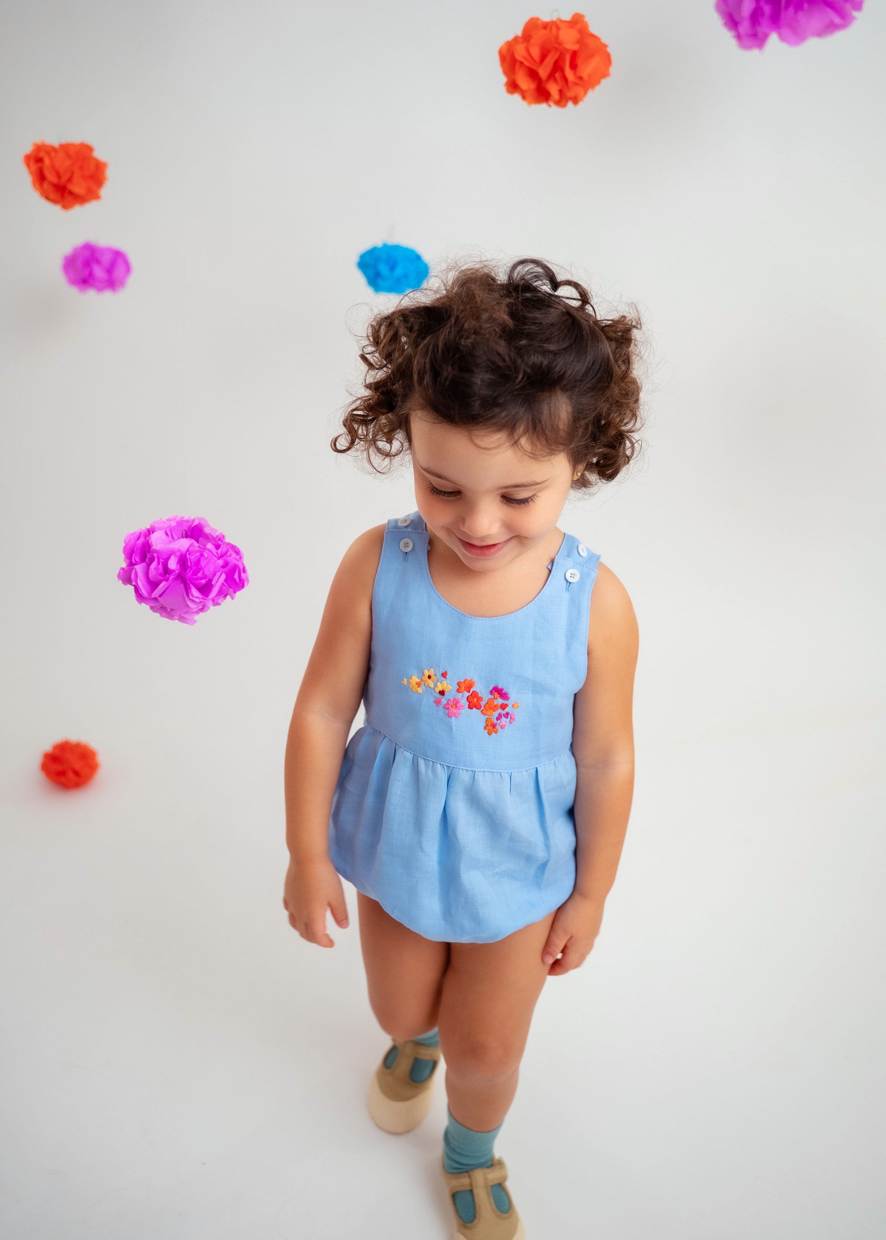 Menina usando macaquinho estilo romper de bebê em linho azul-celeste com bordado de flores.