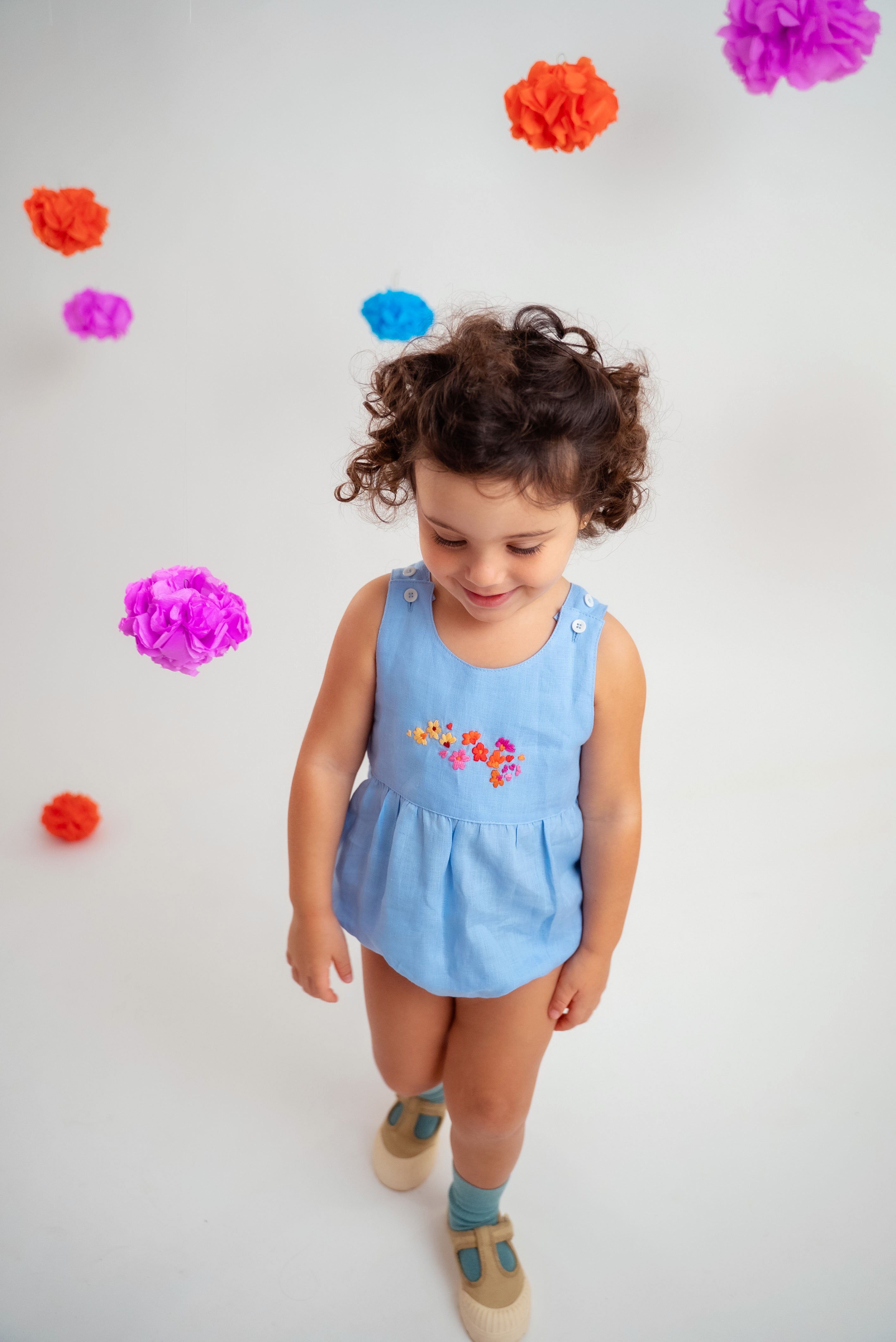 Menina usando macaquinho estilo romper de bebê em linho azul-celeste com bordado de flores.