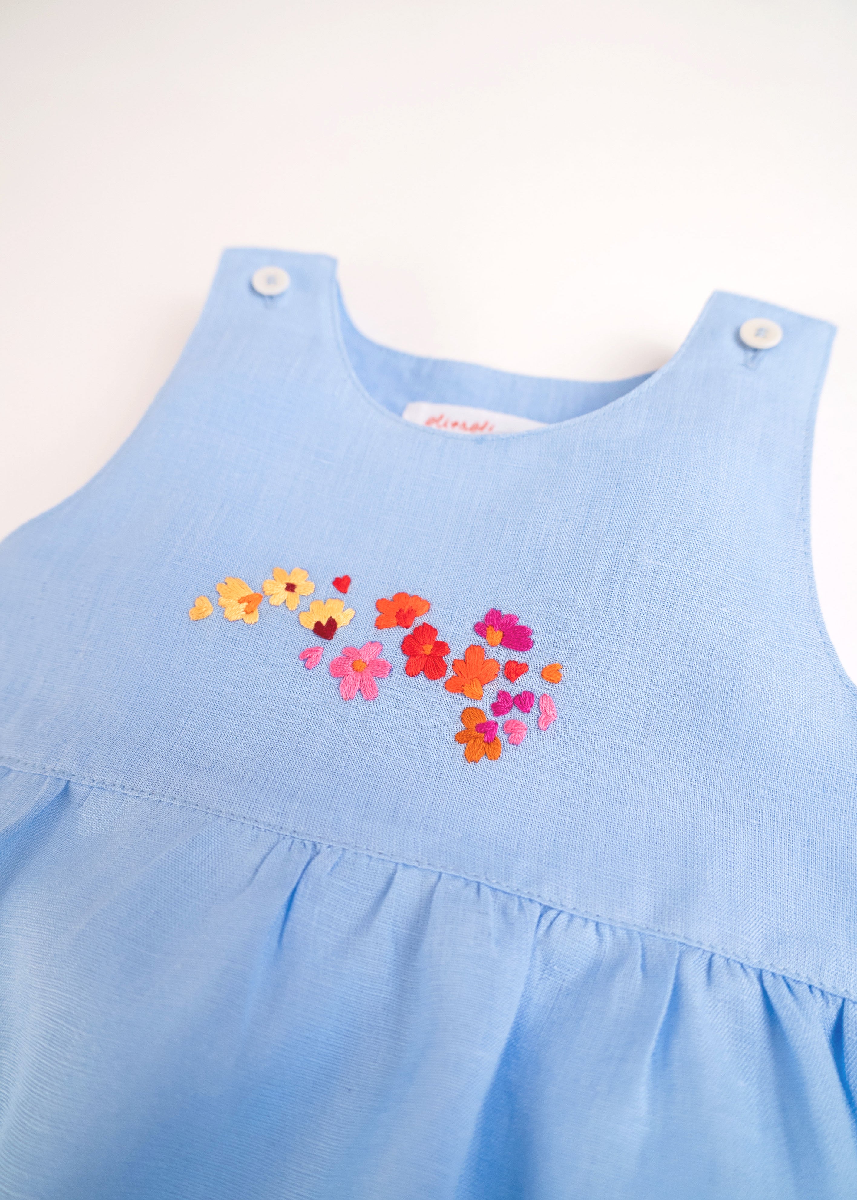 Detalhe do bordado à mão de flores do macaquinho de bebê menina em puro linho azul.