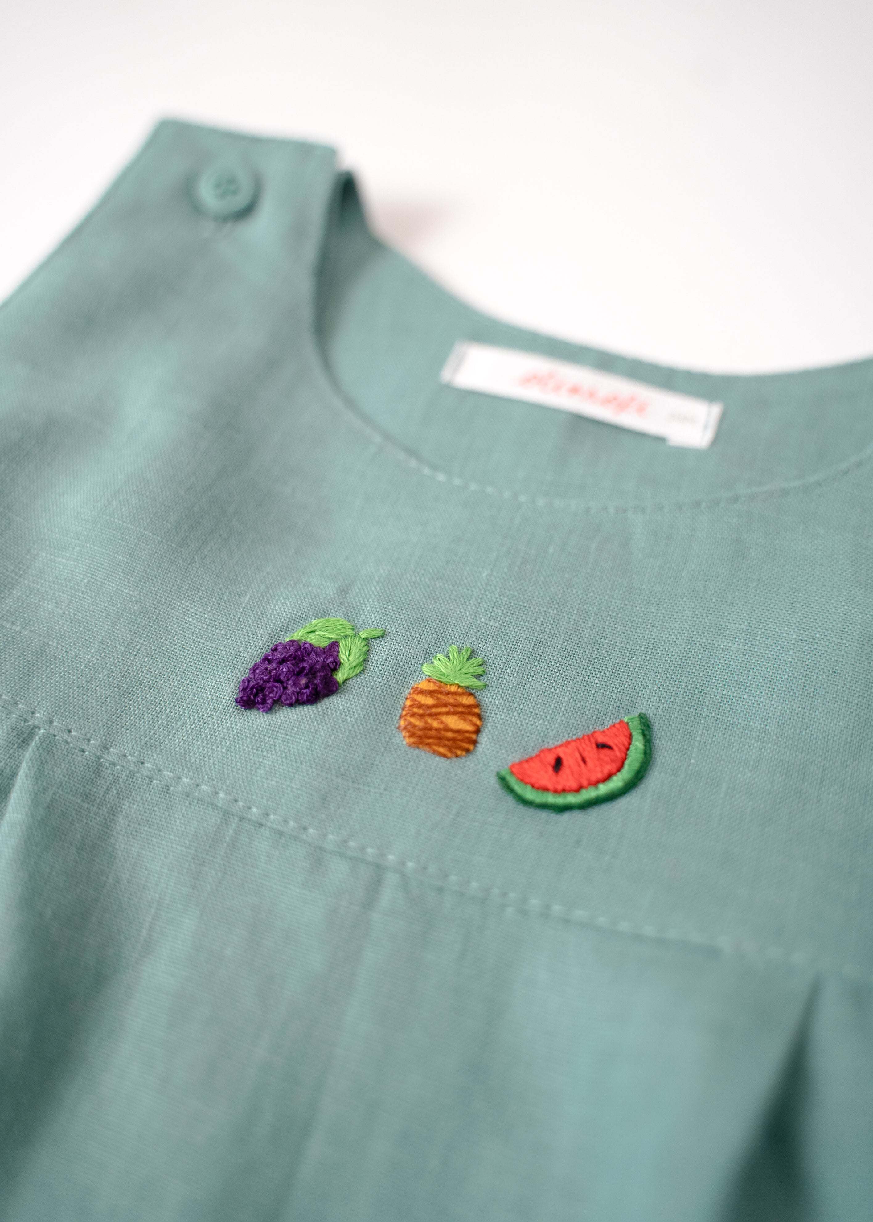 Close do peitinho do macaquinho de linho azul laguna com bordado à mão de uva, abacaxi e melancia.