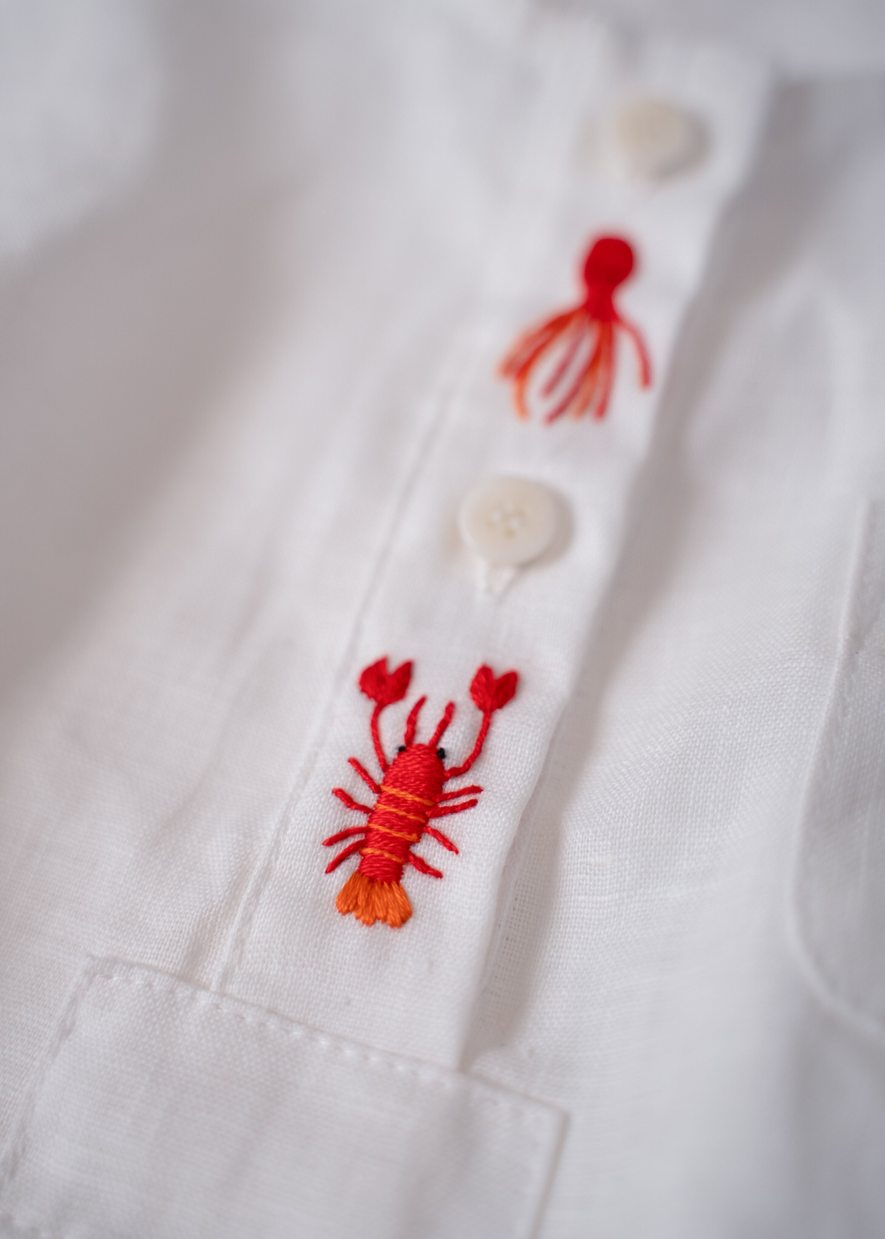 Detalhe do bordado à mão de lagosta vermelha em macaquinho de bebê em linho branco.