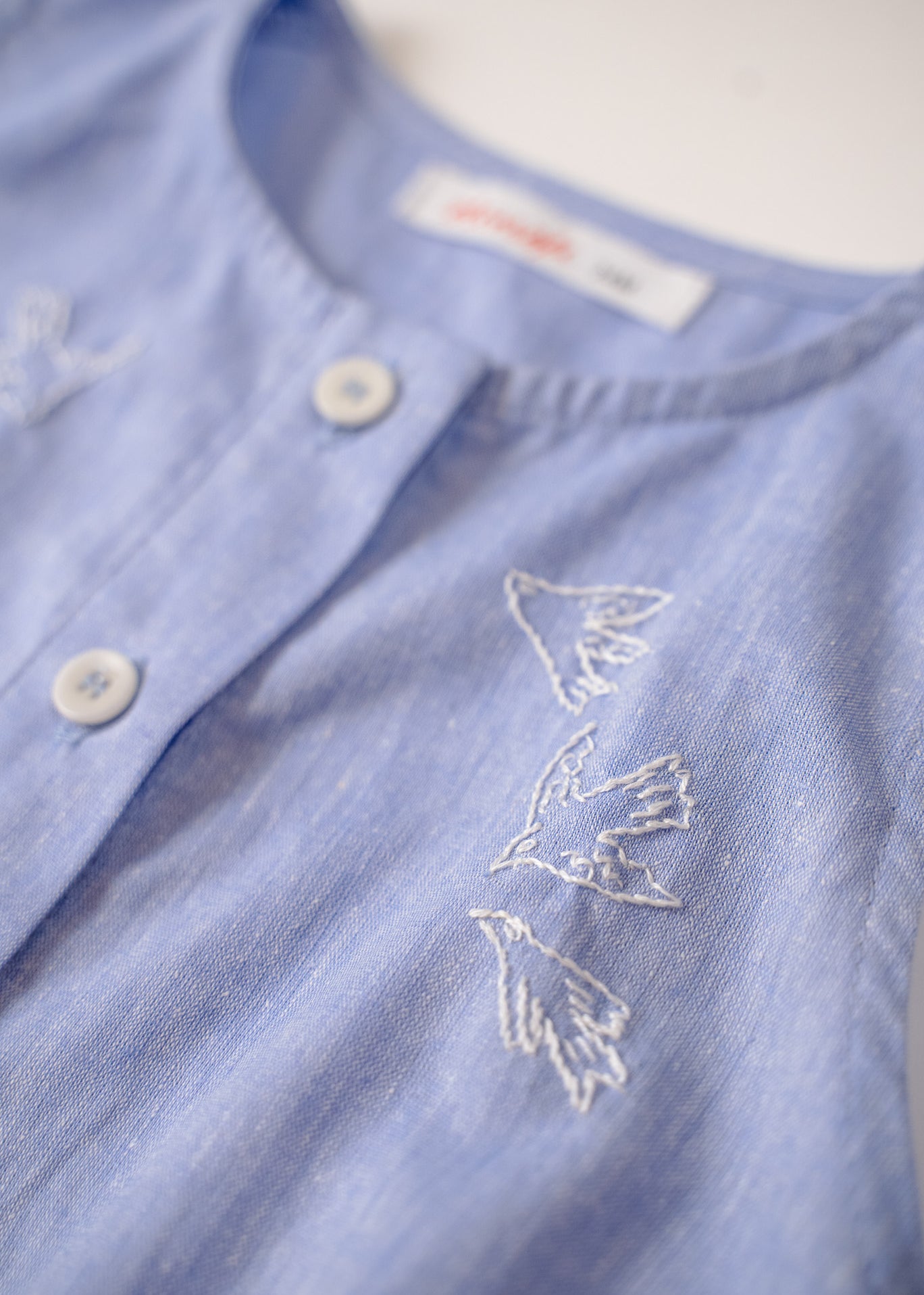 Detalhe do bordado à mão de pássaros brancos do macaquinho de bebê em linho misto azul.