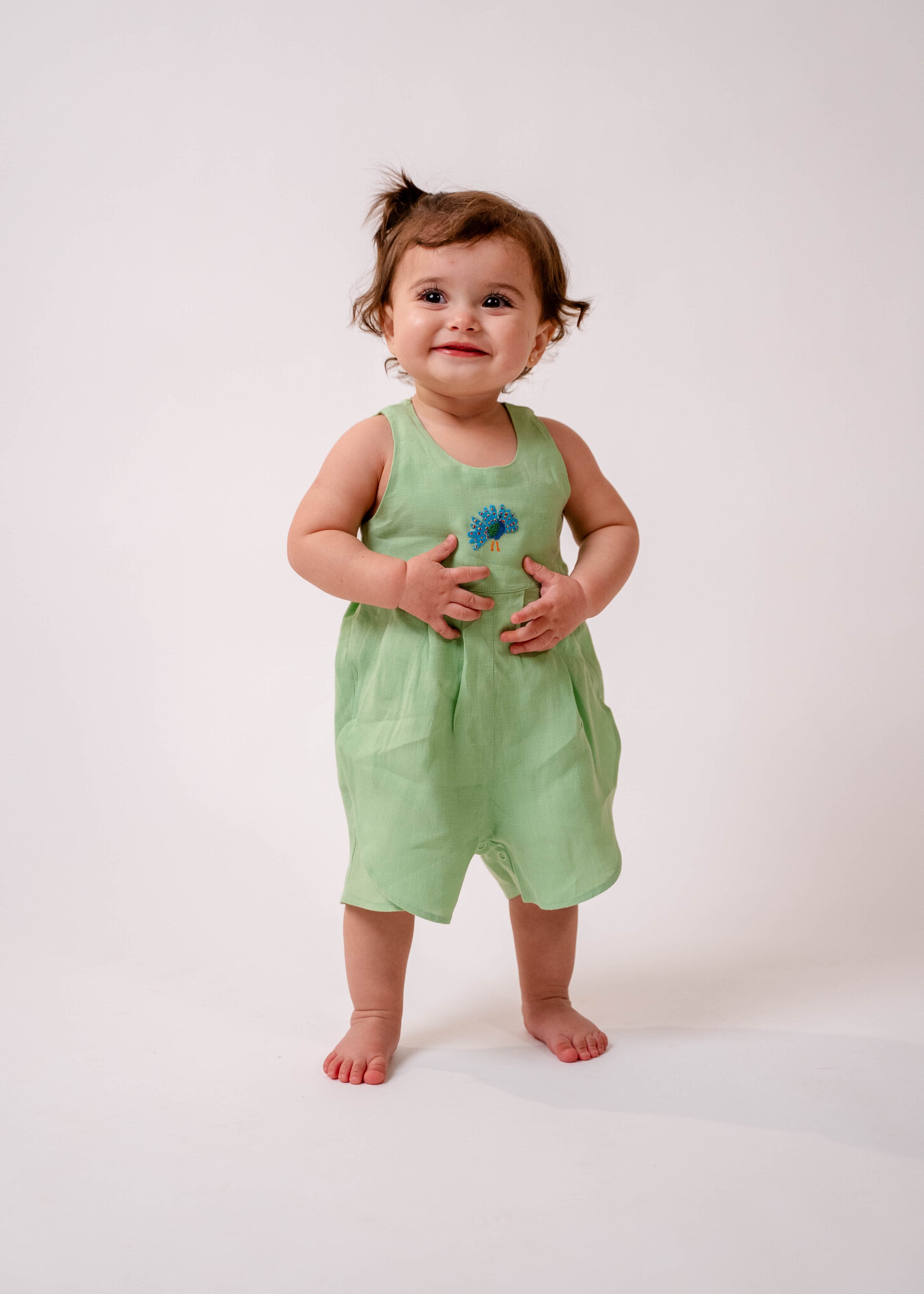 Bebê menina usando macaquinho em linho puro verde claro com bordado manual de pavão no peito.