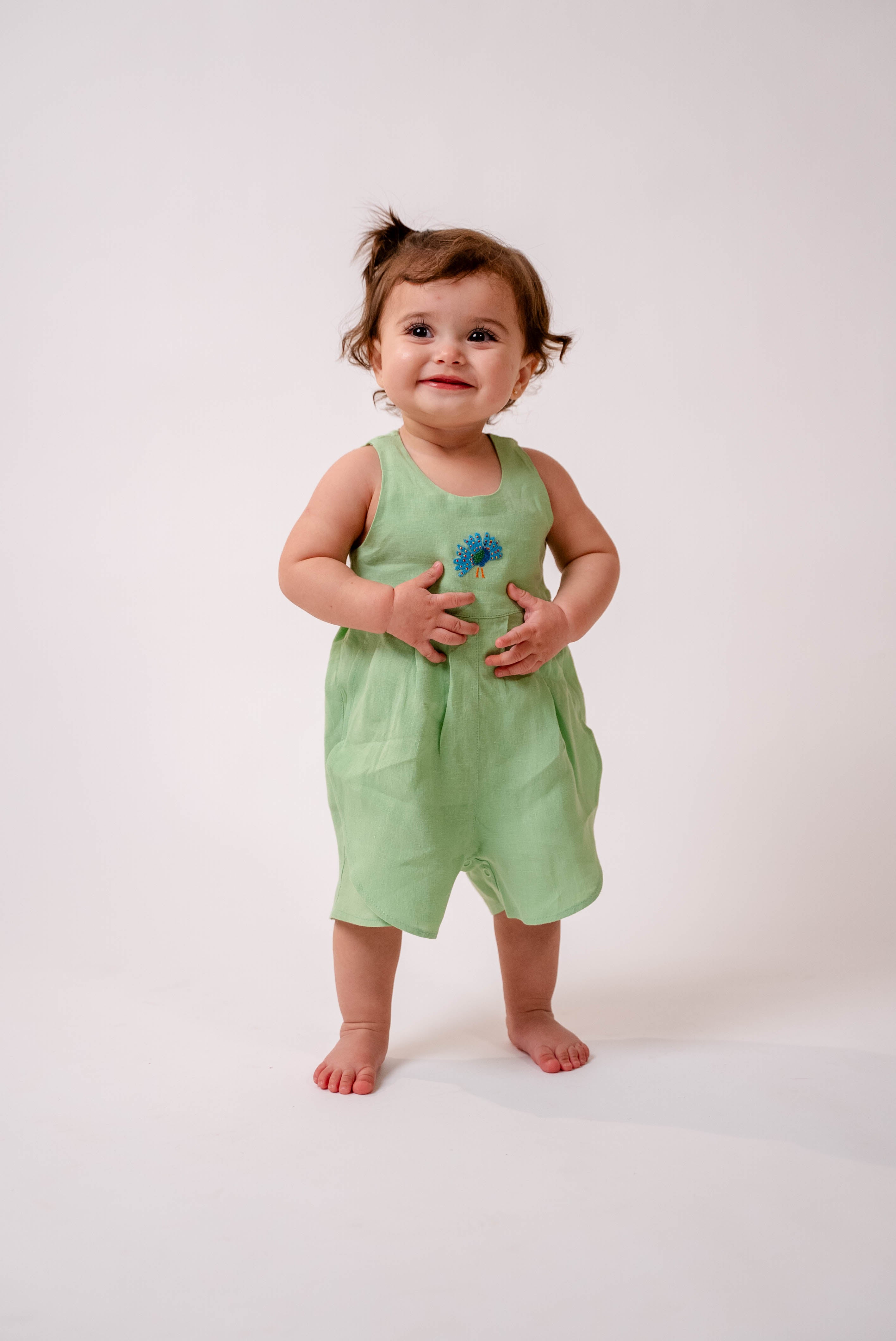 Bebê menina usando macaquinho em linho puro verde claro com bordado manual de pavão no peito.