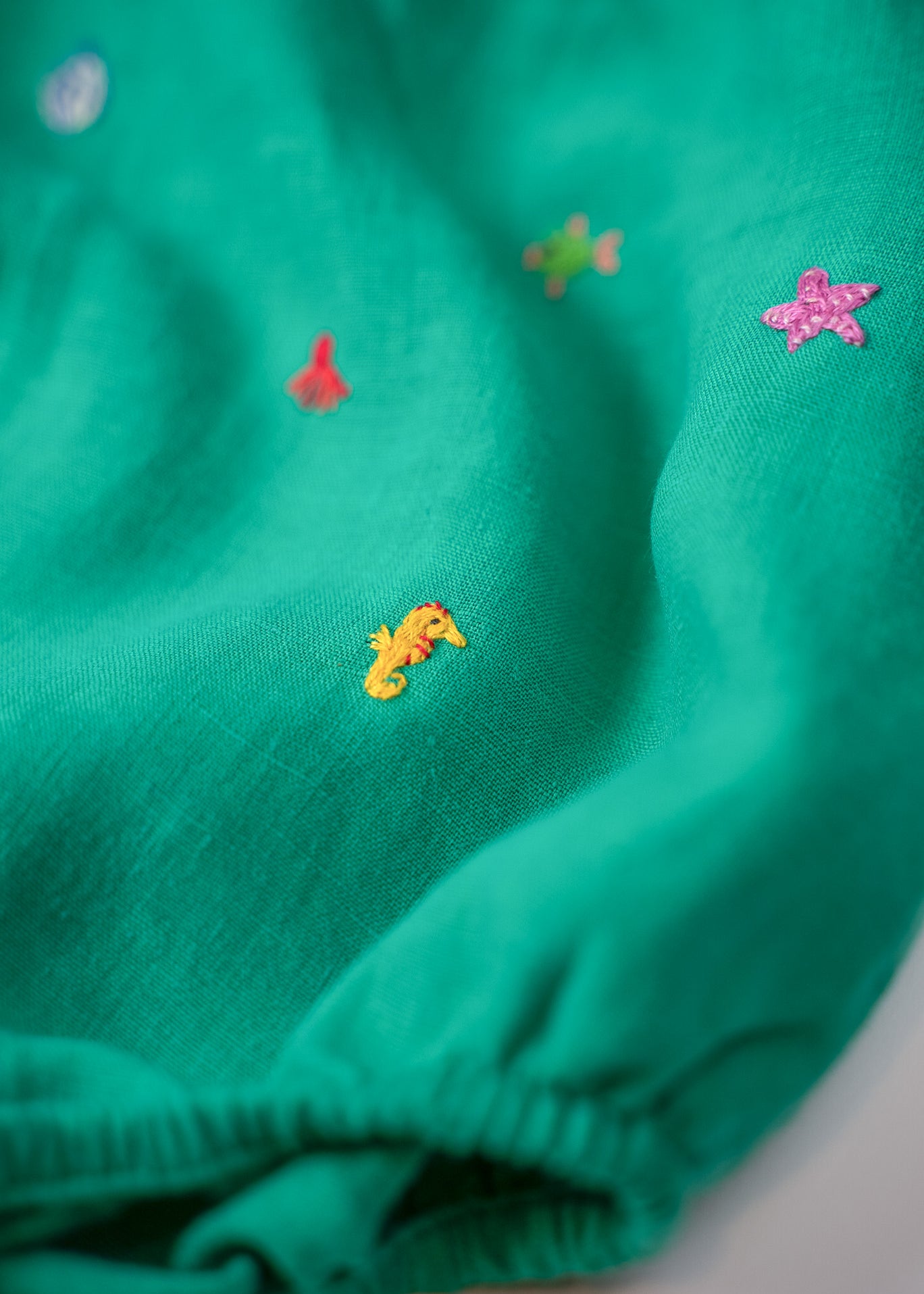 Close dos bordados à mão de cavalo-marinho amarelo e estrela-do-mar rosa nas perninhas do macaquinho de bebê.