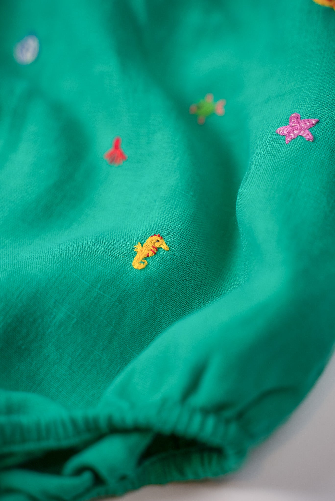 Close dos bordados à mão de cavalo-marinho amarelo e estrela-do-mar rosa nas perninhas do macaquinho de bebê.