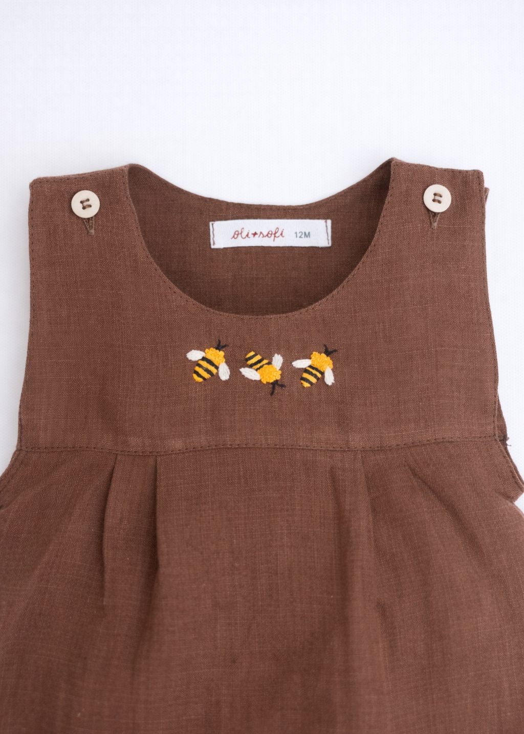 Macaquinho de bebê em linho marrom com três abelhas bordadas à mão no peito, visto em detalhe.
