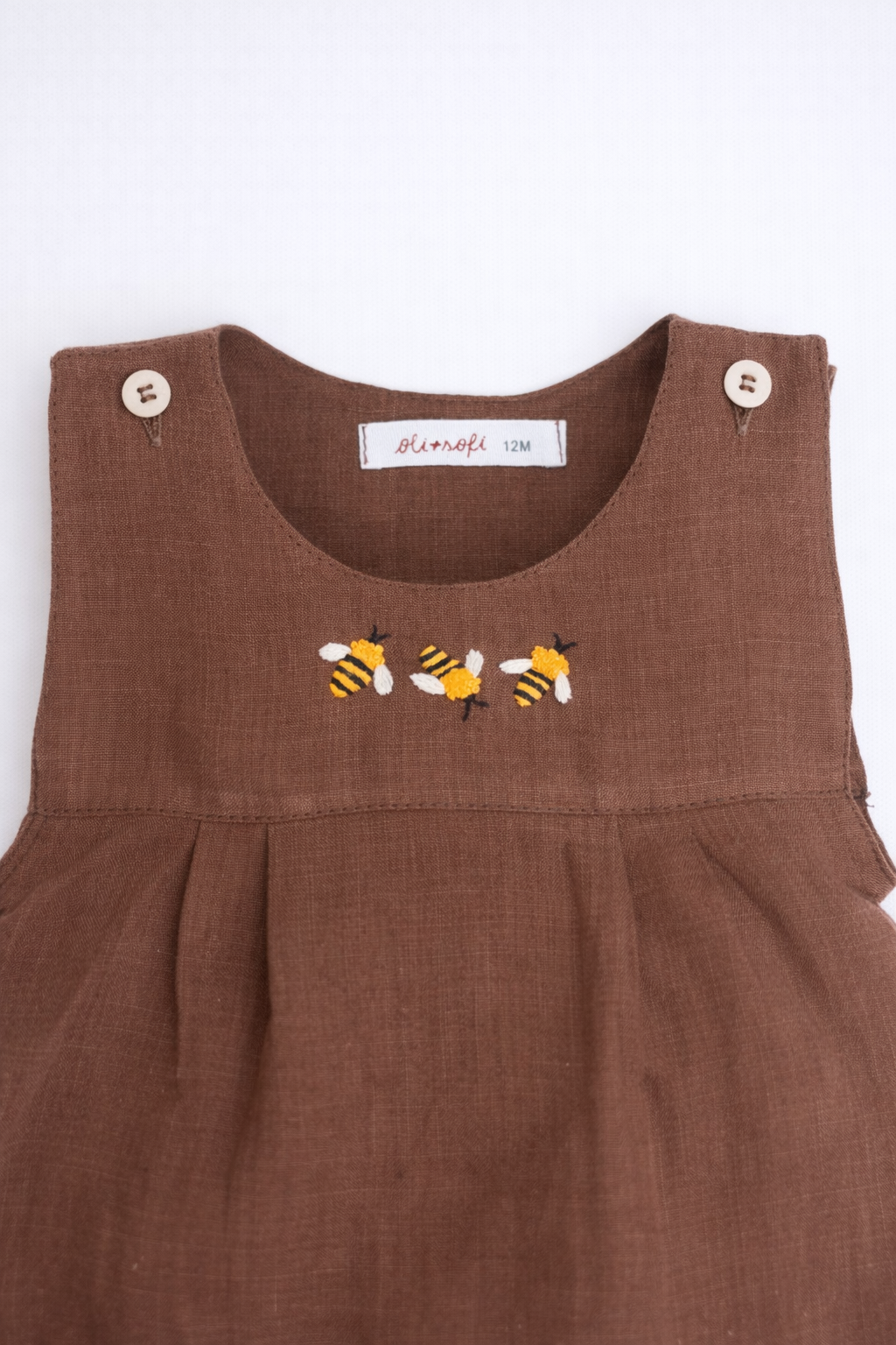 Macaquinho de bebê em linho marrom com três abelhas bordadas à mão no peito, visto em detalhe.