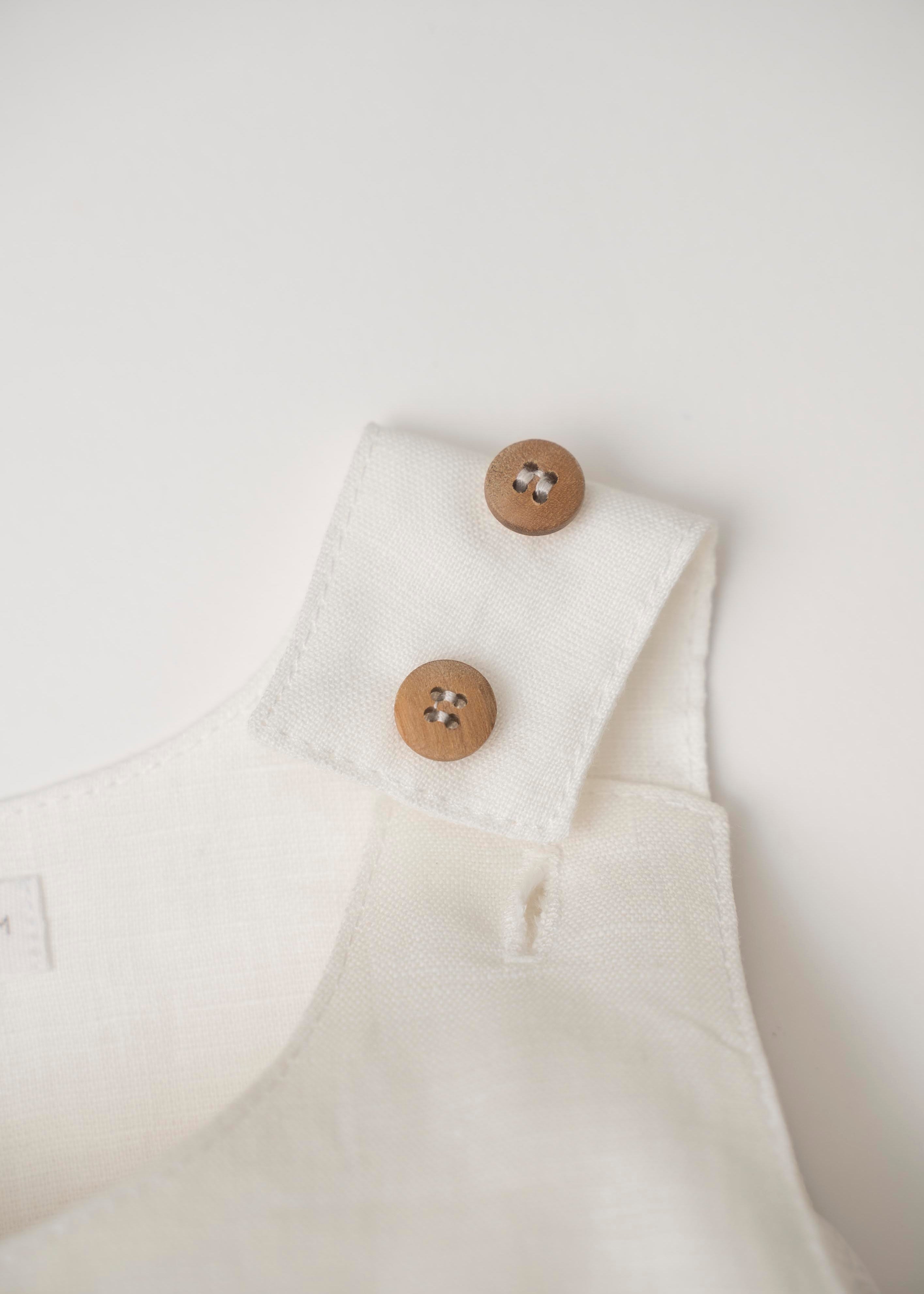 Detalhe do fechamento no ombro com botões de madeira e duas regulagens de altura do macaquinho de bebê em linho branco.