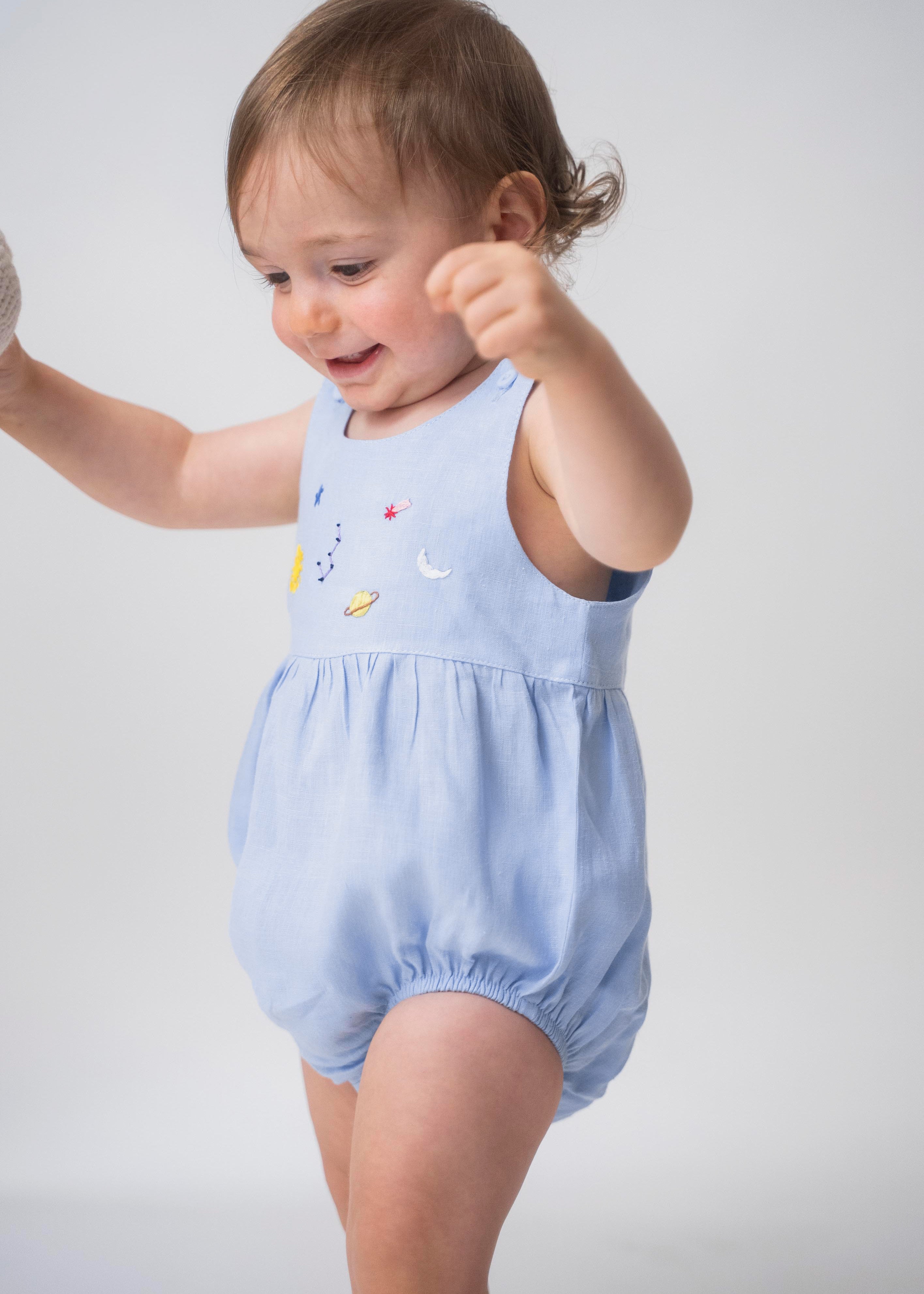 Bebê usando macaquinho de linho azul claro com modelagem balonê e bordado artesanal de sol, lua e estrelas.