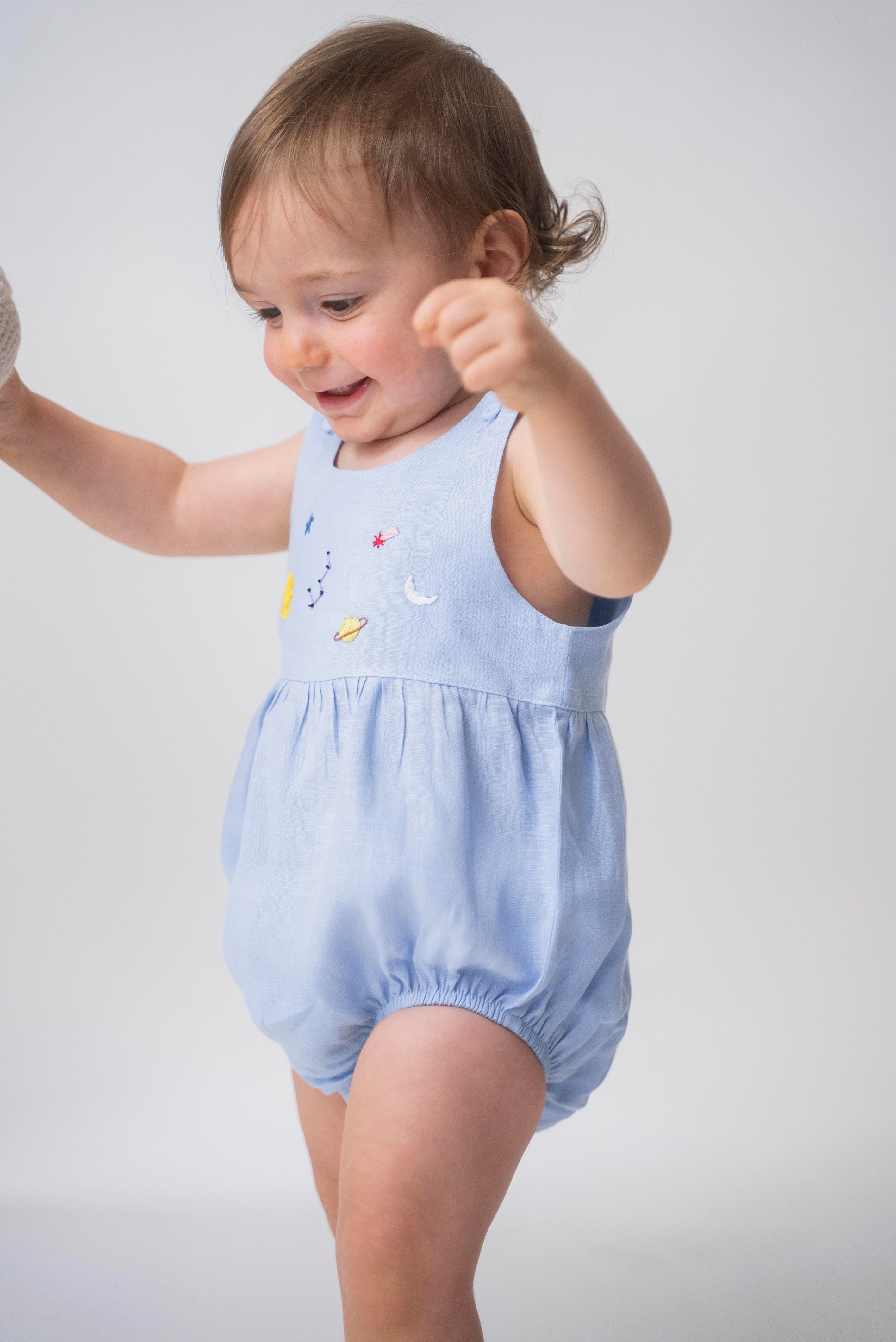 Bebê usando macaquinho de linho azul claro com modelagem balonê e bordado artesanal de sol, lua e estrelas.