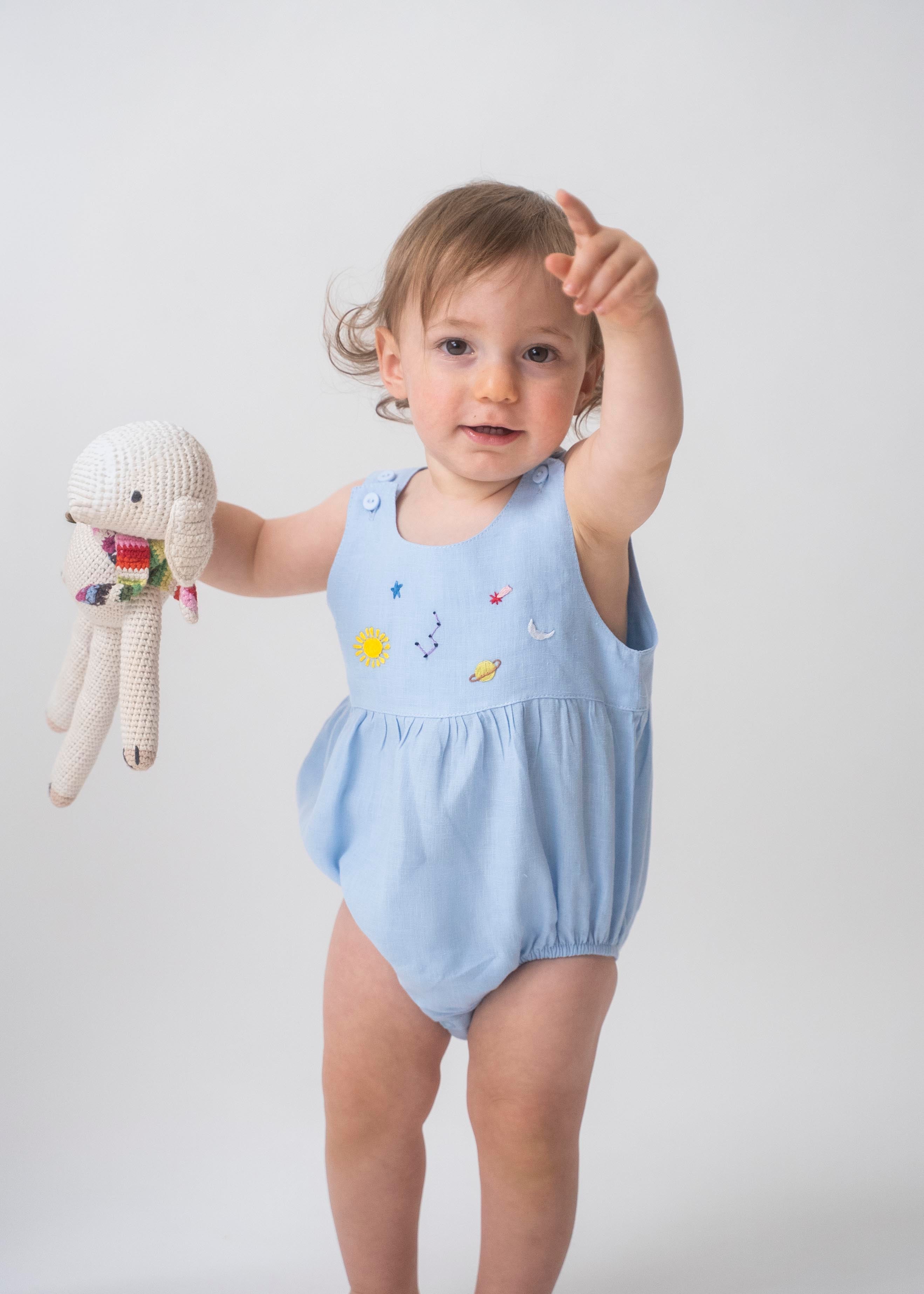Bebê usando macaquinho de linho azul claro com modelagem balonê e bordado artesanal de sol, lua e estrelas.