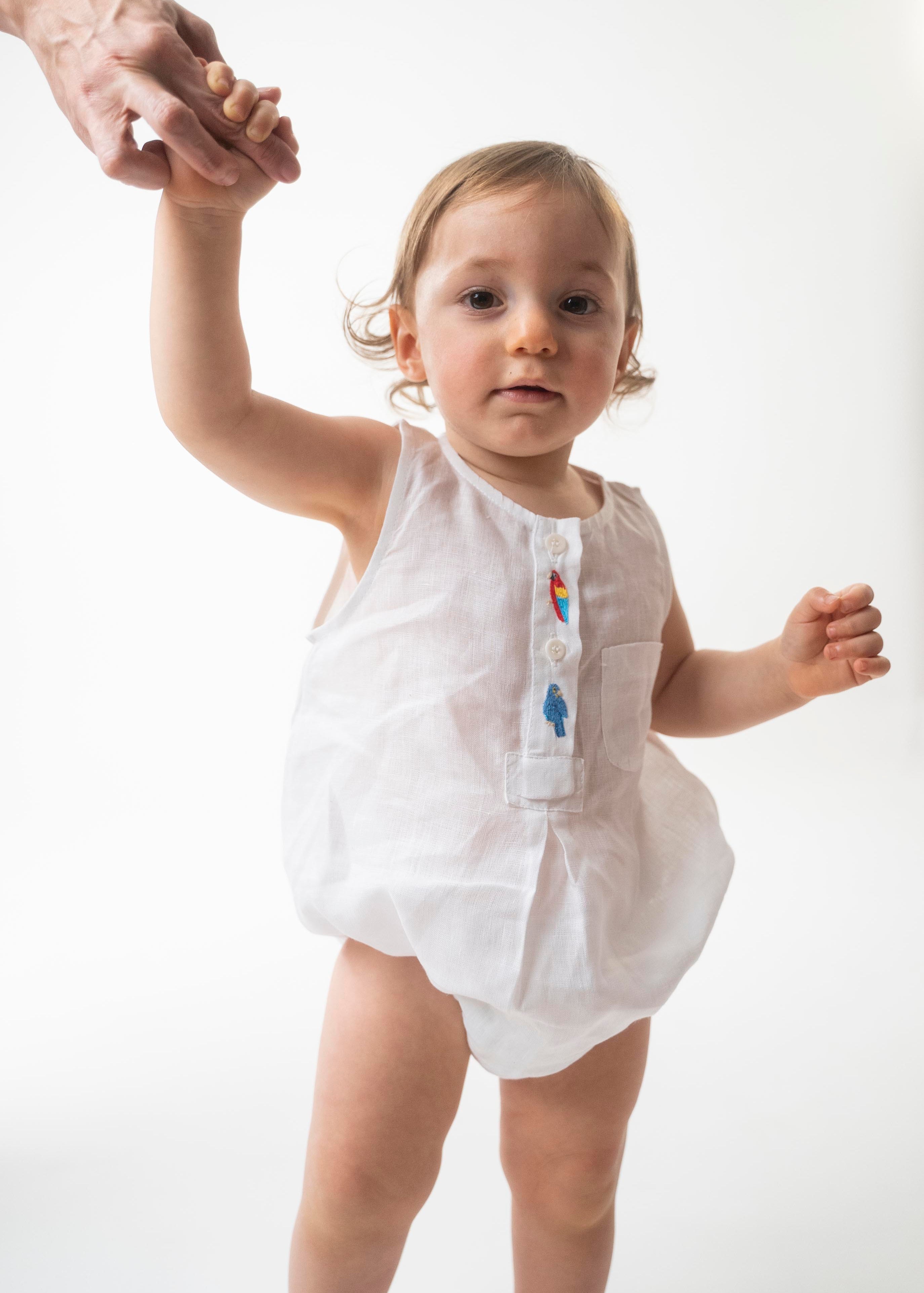 Bebê usando Macaquinho Valen de linho branco com araras bordadas à mão e modelagem soltinha.