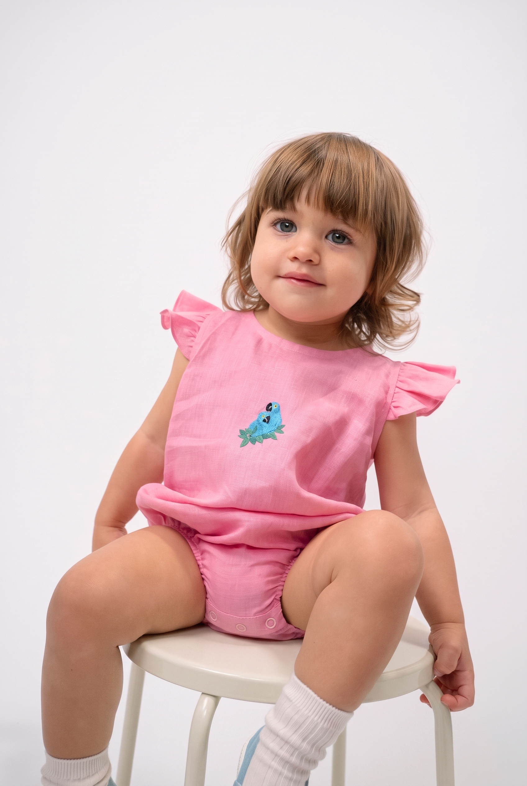 Bebê sentada vestindo Macaquinho Clara em linho rosa com bordado à mão de duas araras azuis sobre folhas verdes.