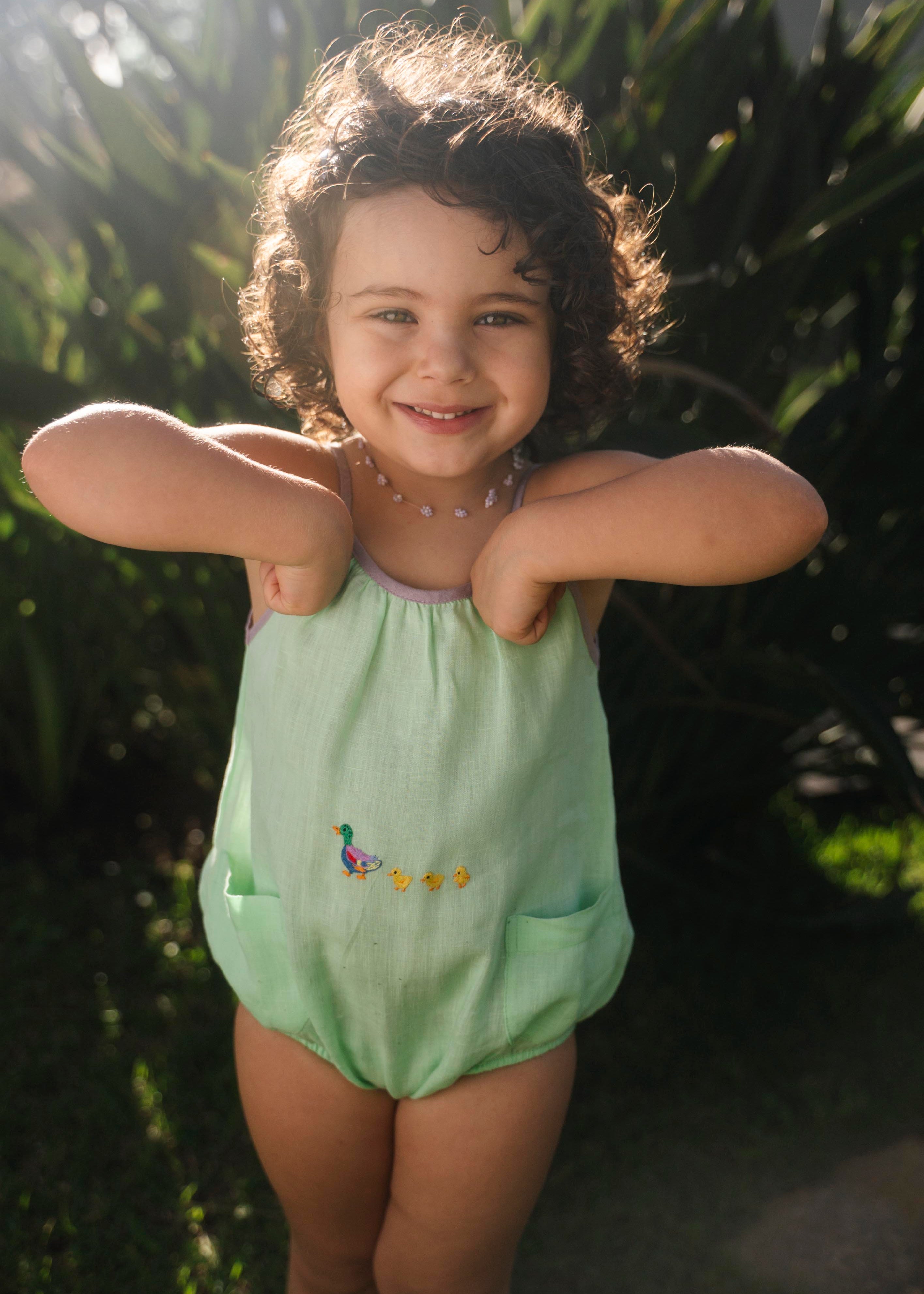 Macaquinho de linho verde menta no corpo, alças com viés lilás e patinhos bordados ao centro.