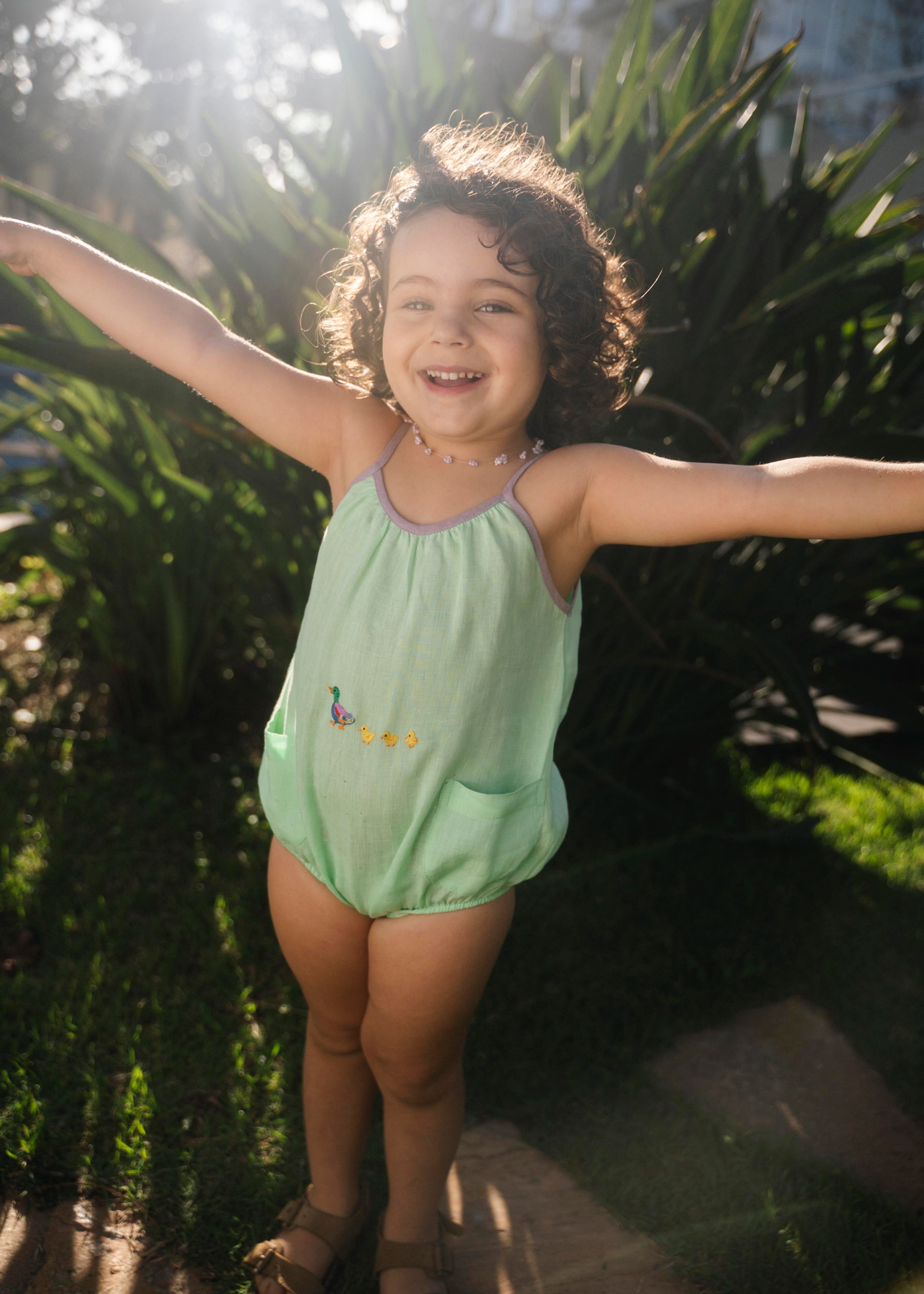 Menina vestindo macaquinho bebê verde menta com patinhos bordados, modelagem ampla com franzido delicado e elástico nas perninhas.