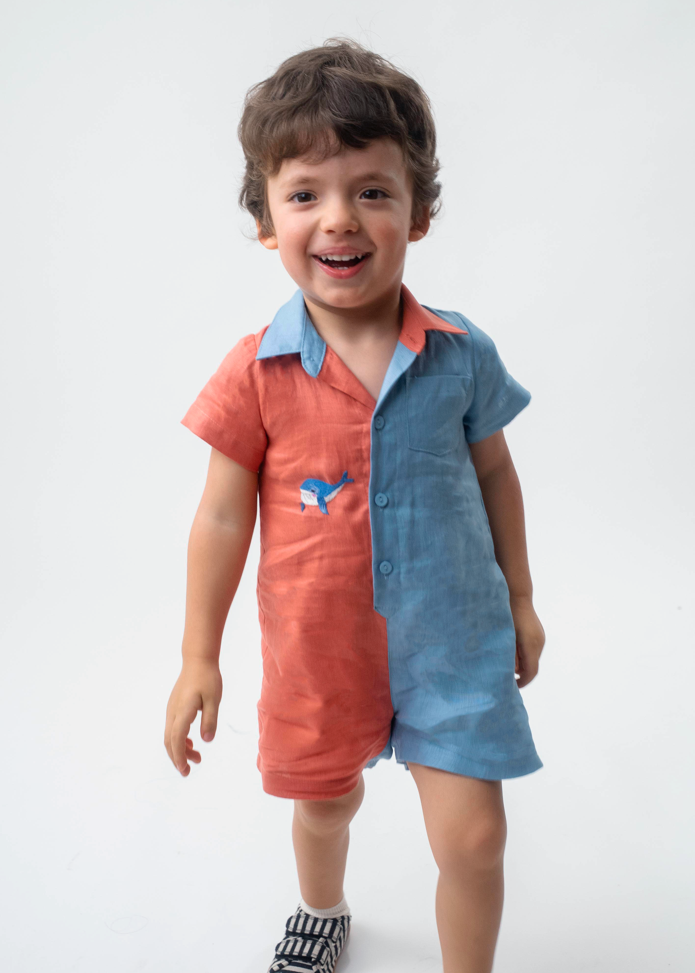 Menino vestindo Macaquinho Tiê infantil em linho coral e azul celeste com bordado à mão de baleia.