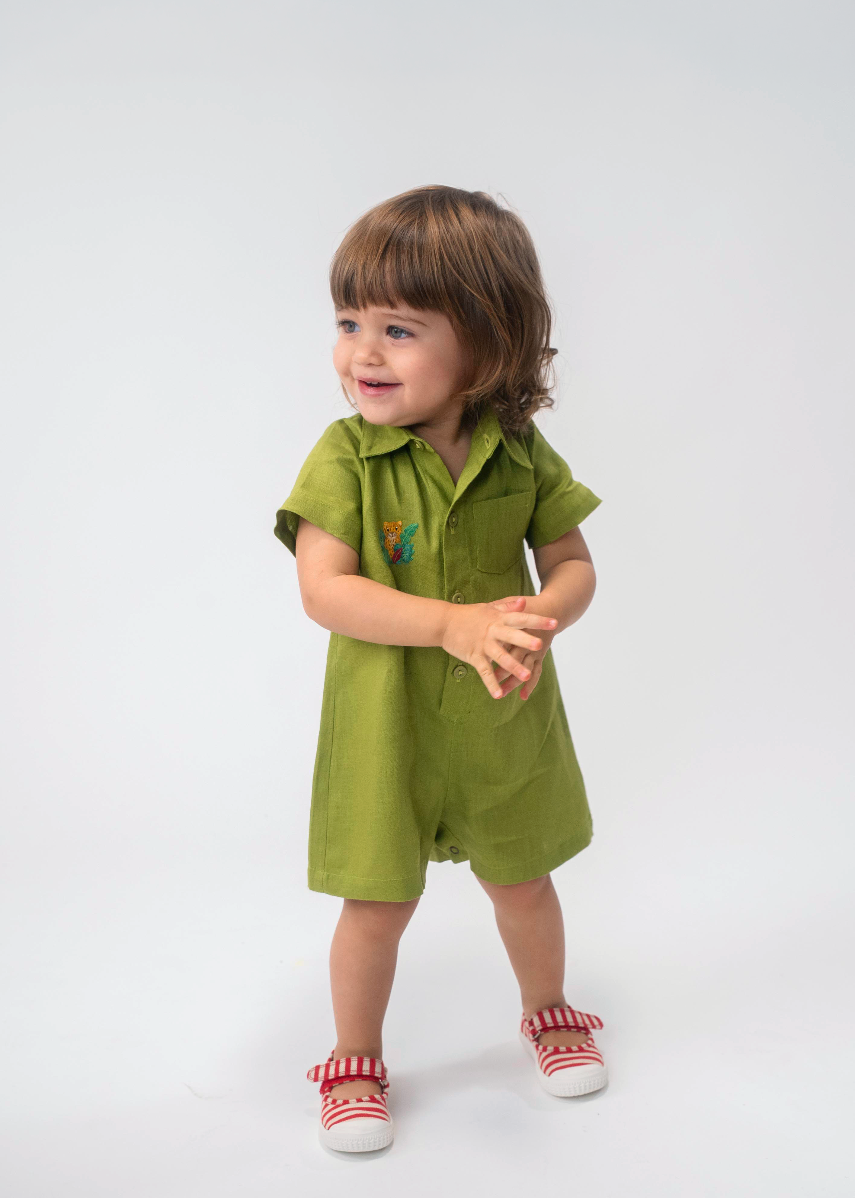 Criança usando Macaquinho Tiê infantil em linho verde com gola, mangas curtas e bordado de onça à mão.