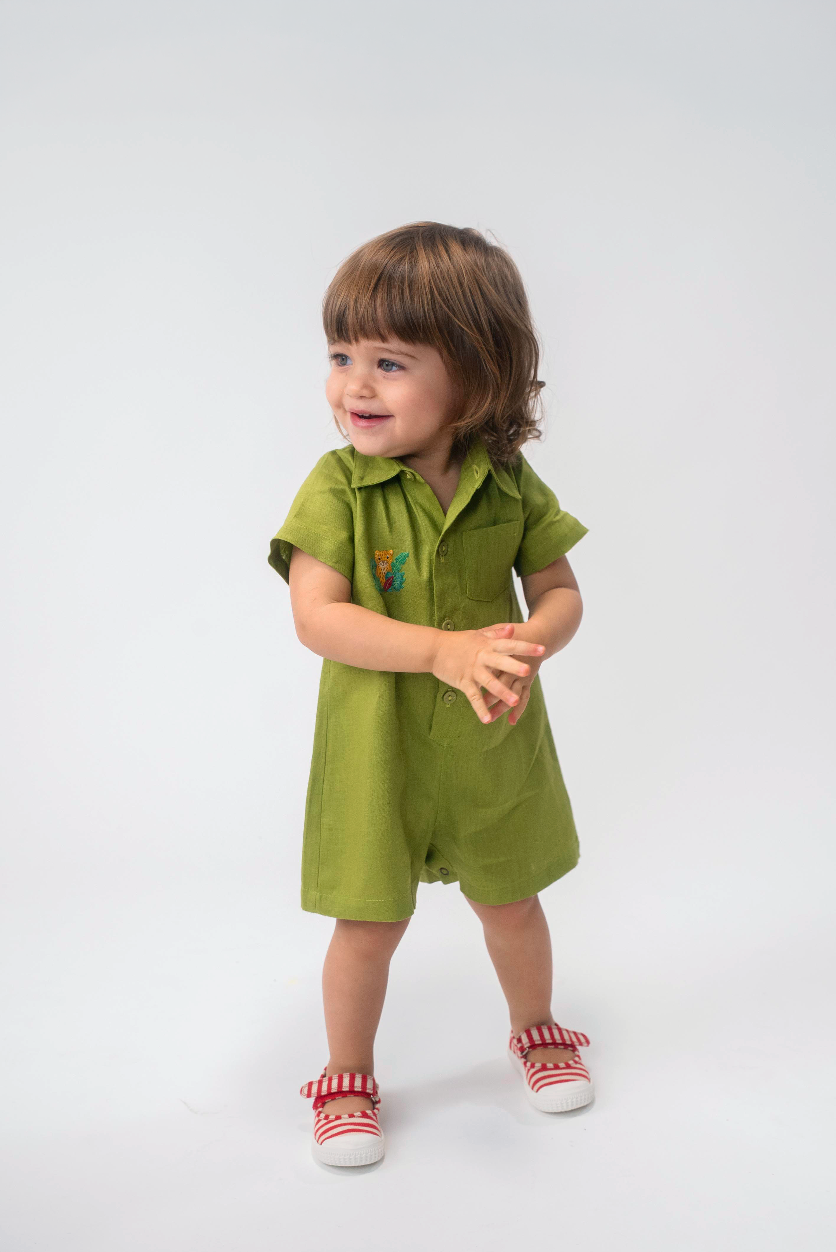 Criança usando Macaquinho Tiê infantil em linho verde com gola, mangas curtas e bordado de onça à mão.