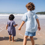 Macaquinho de linho azul jeans no corpo, com gola violeta e modelagem curta, visto de costas em menino na praia.