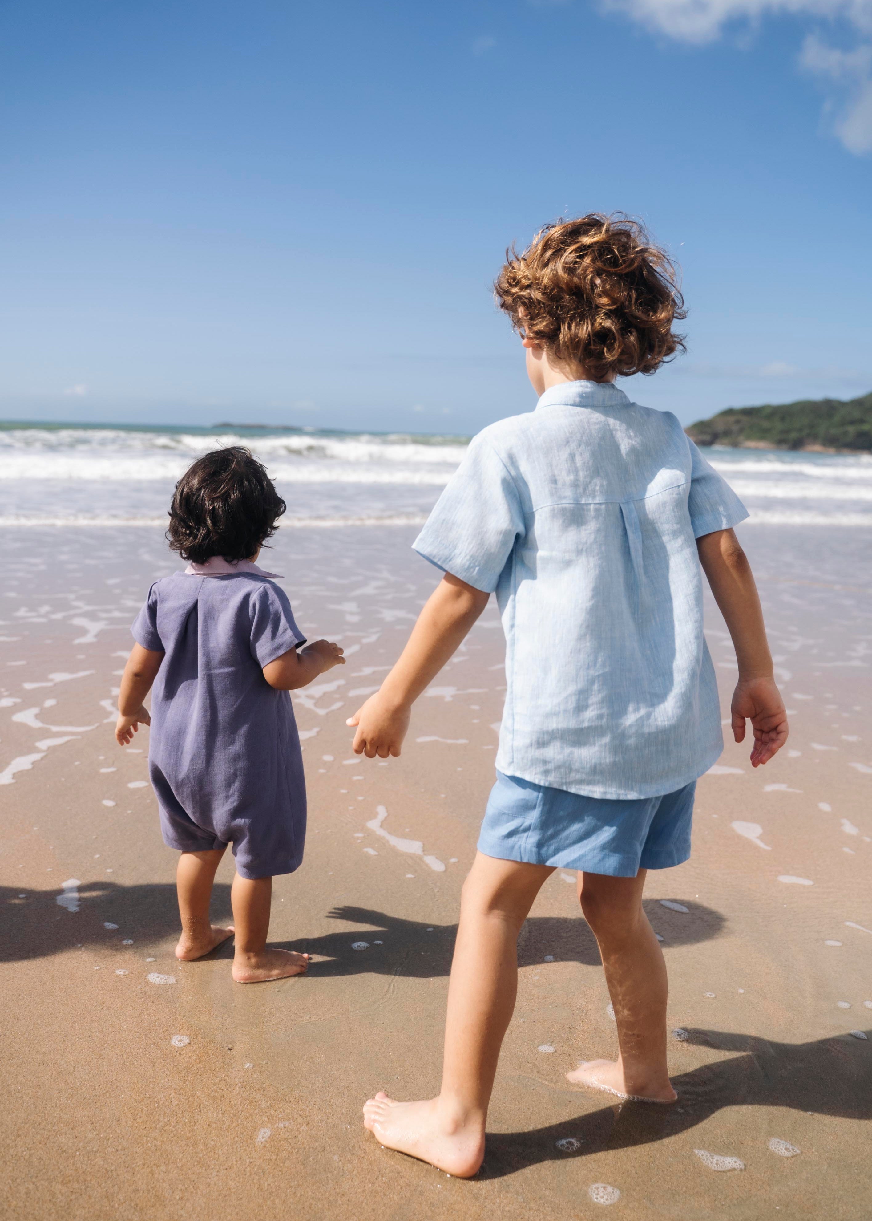 Macaquinho de linho azul jeans no corpo, com gola violeta e modelagem curta, visto de costas em menino na praia.