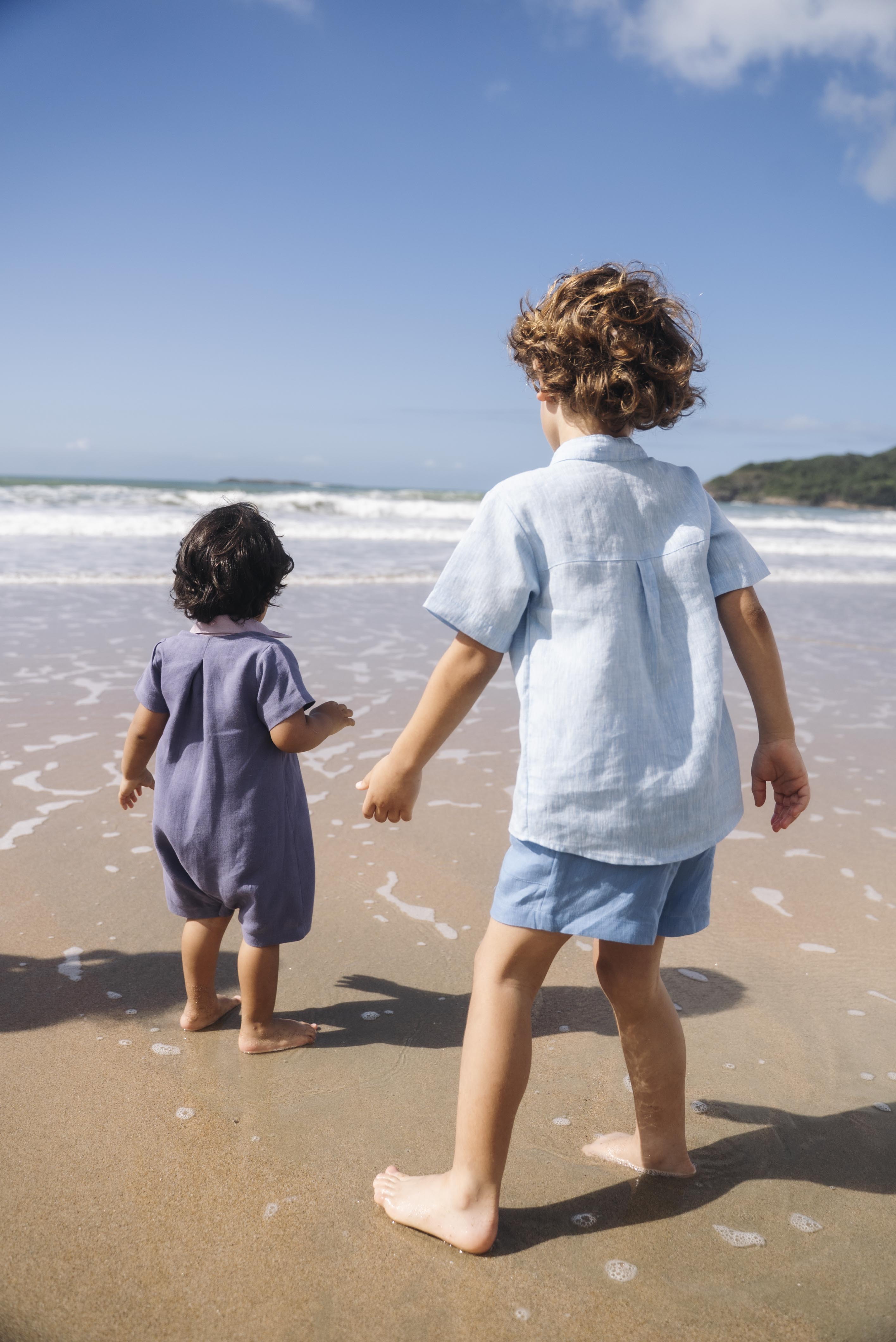 Macaquinho de linho azul jeans no corpo, com gola violeta e modelagem curta, visto de costas em menino na praia.