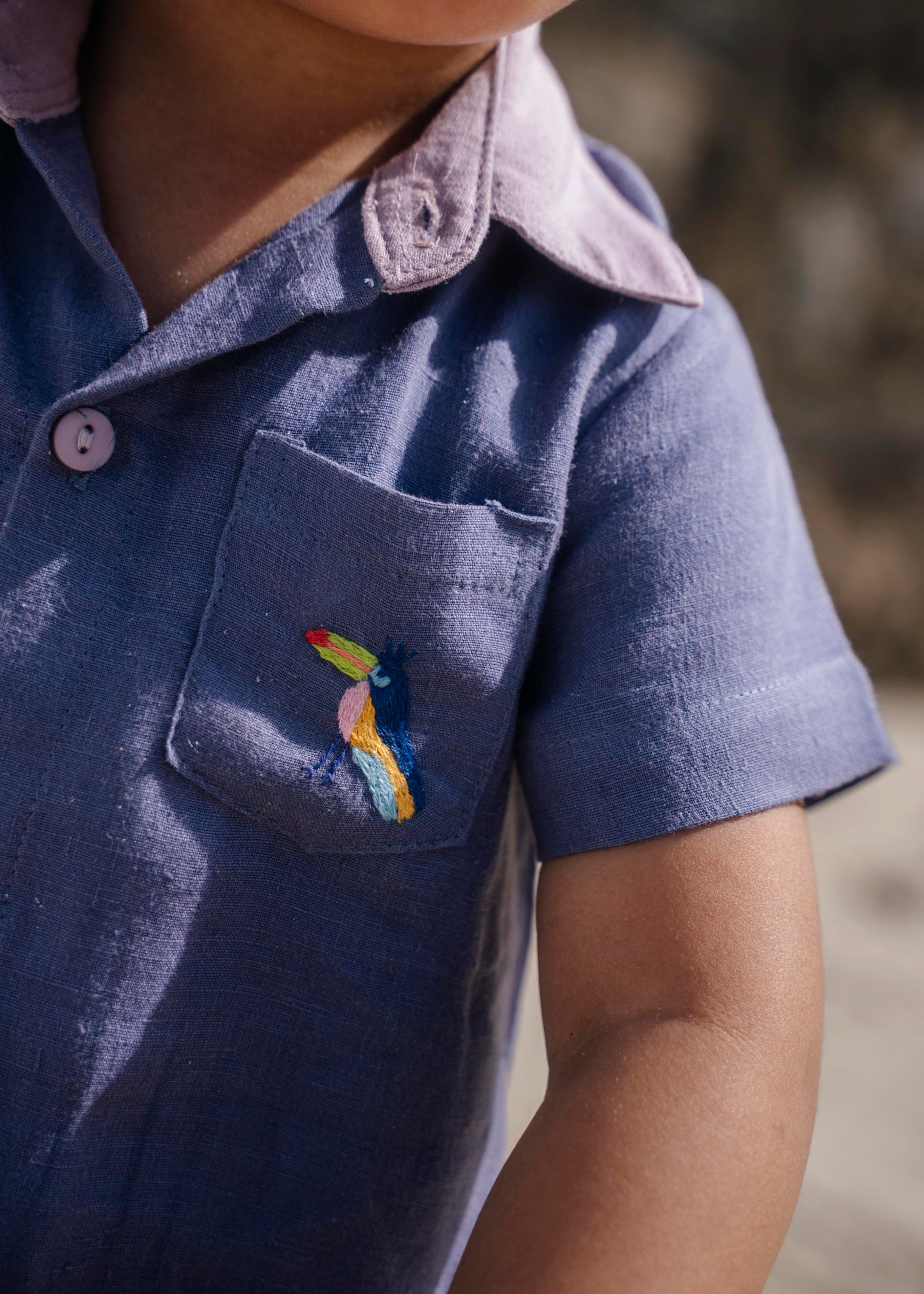 Detalhe do tucano bordado à mão no bolso do macaquinho em linho azul, com gola e botões em tom de violeta.