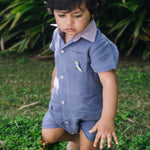 Menino com macaquinho infantil de linho azul jeans no corpo, gola rosa, bolso com tucano bordado e abertura frontal com botões.