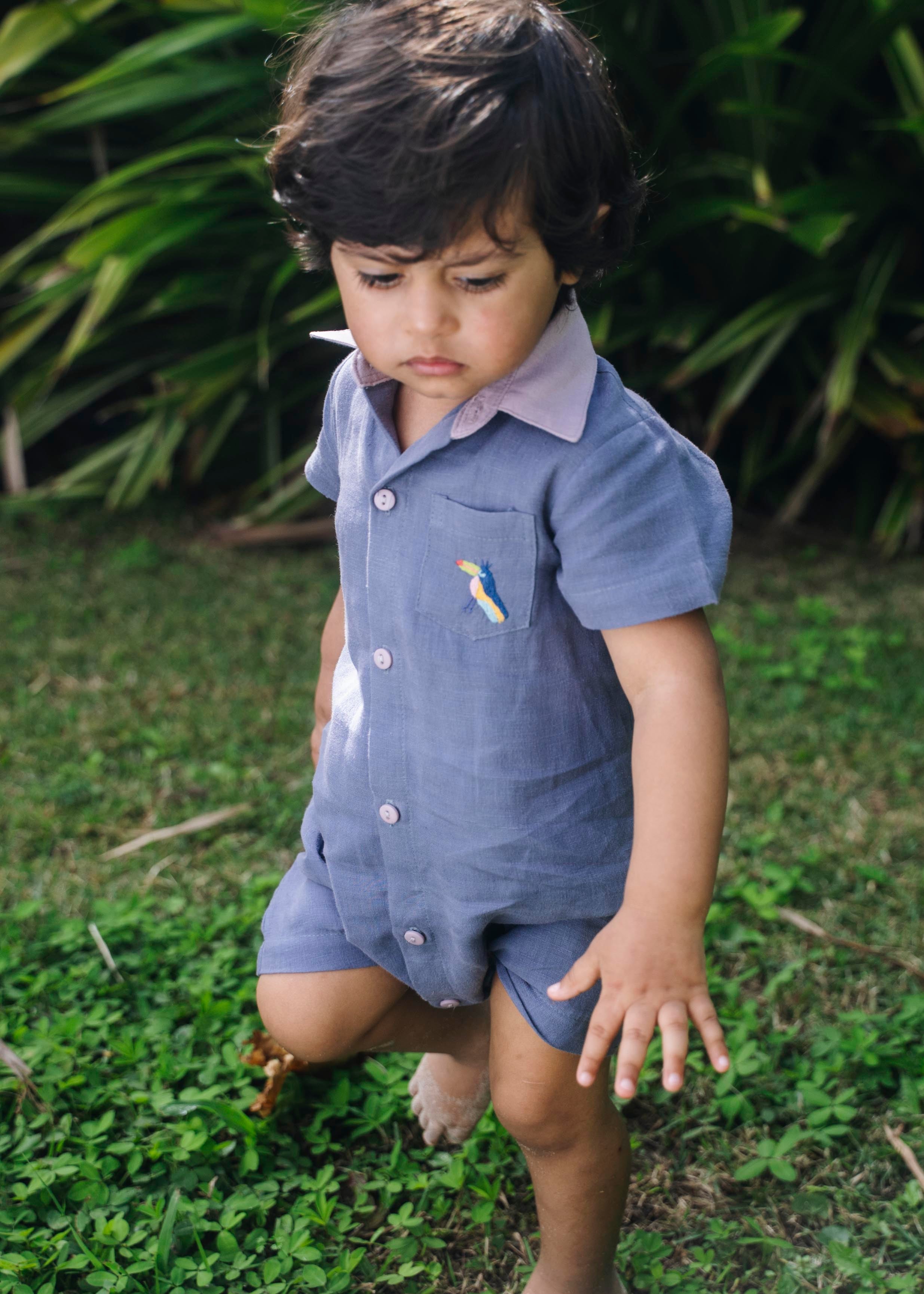 Menino com macaquinho infantil de linho azul jeans no corpo, gola rosa, bolso com tucano bordado e abertura frontal com botões.
