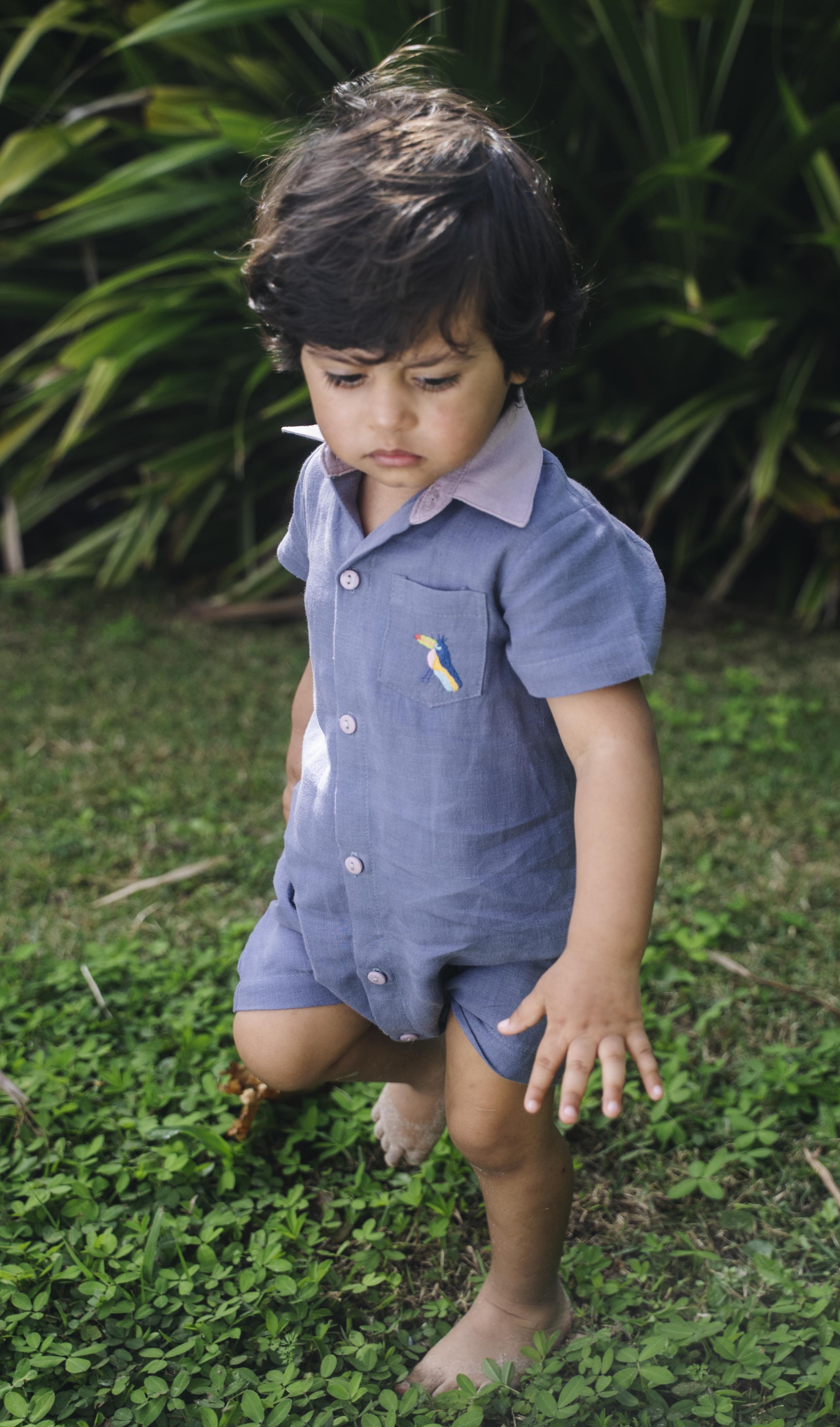 Menino com macaquinho infantil de linho azul jeans no corpo, gola rosa, bolso com tucano bordado e abertura frontal com botões.