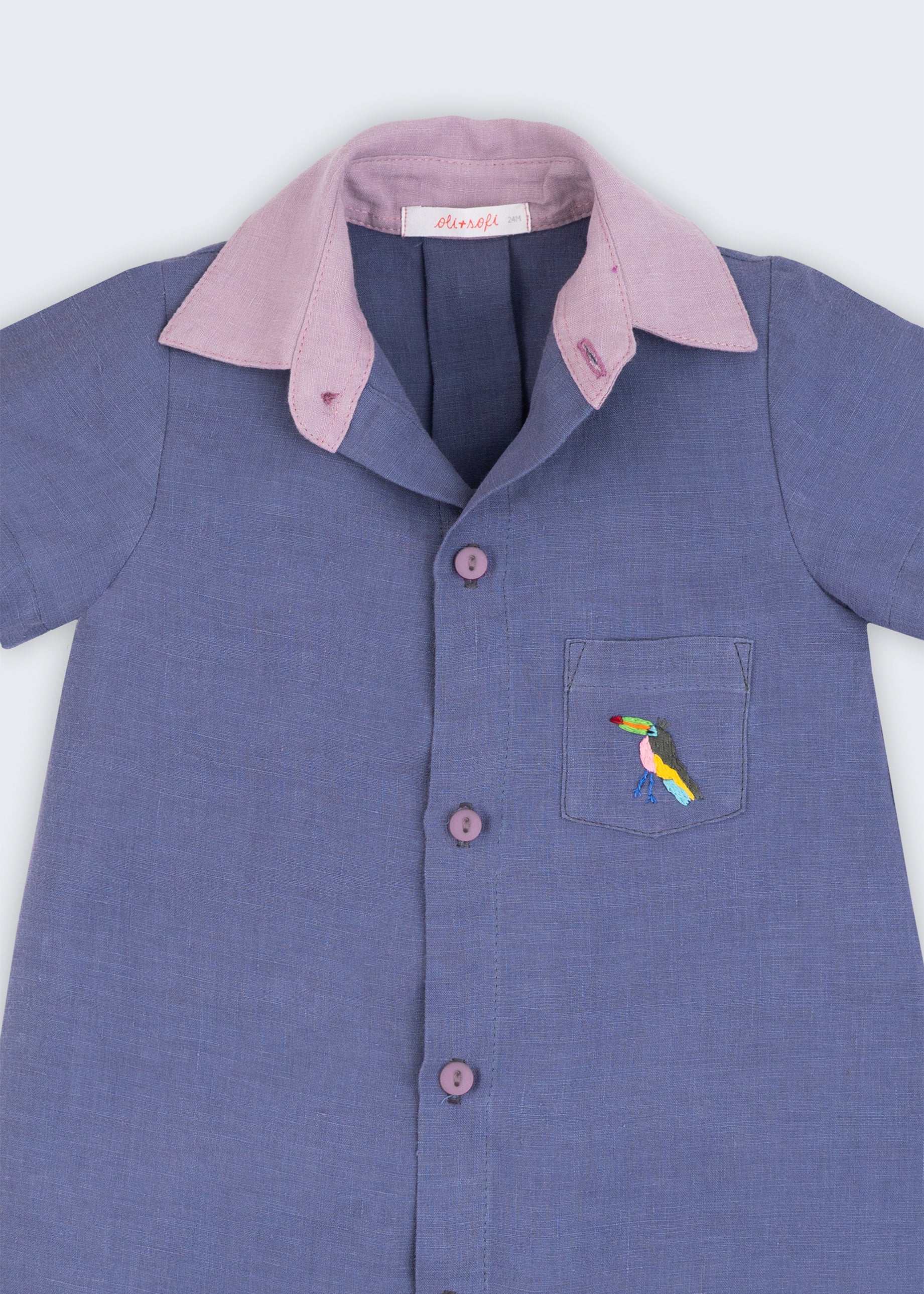 Macaquinho infantil em linho azul jeans com gola violeta e tucano bordado à mão no bolso, visto de frente em detalhe.