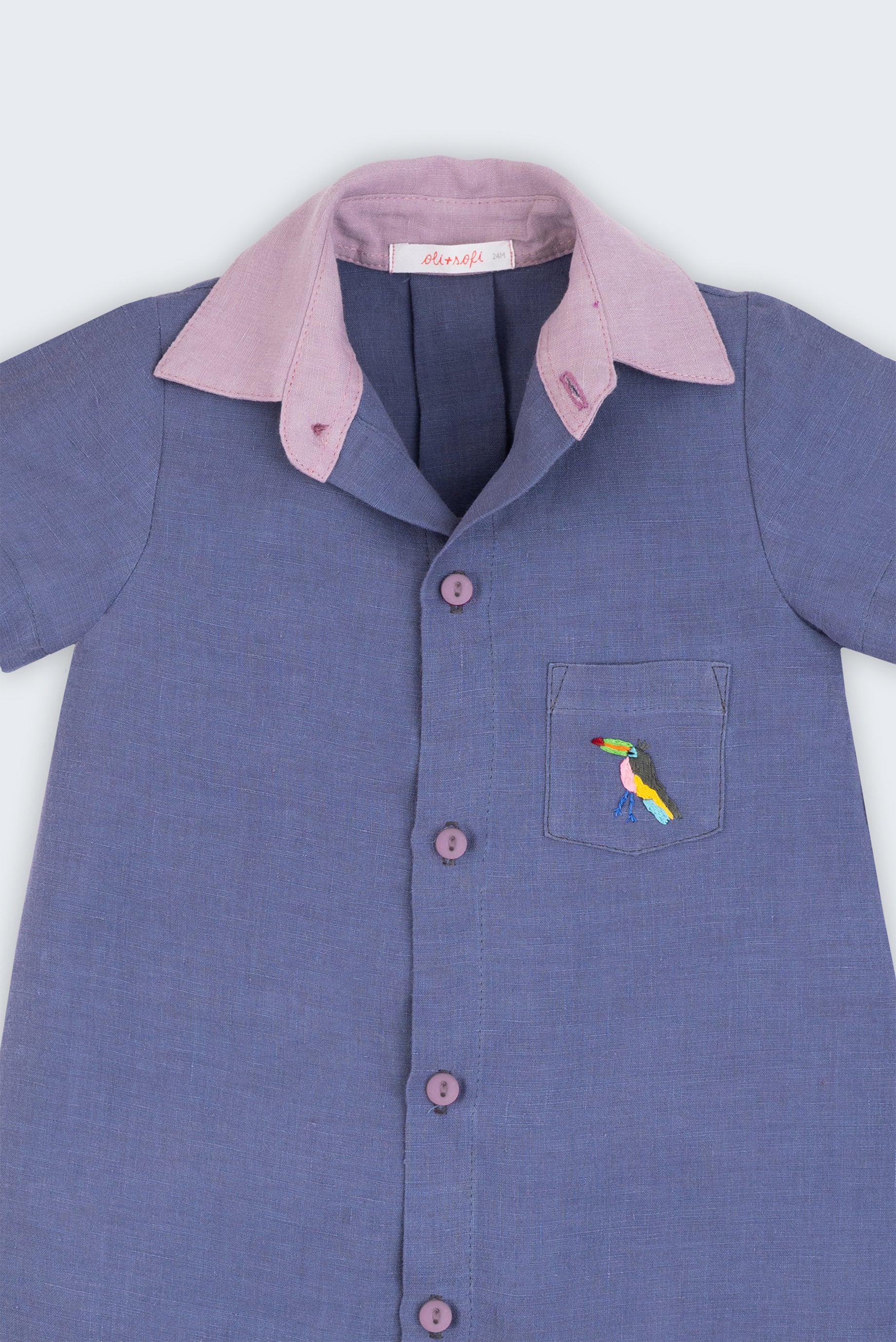 Macaquinho infantil em linho azul jeans com gola violeta e tucano bordado à mão no bolso, visto de frente em detalhe.