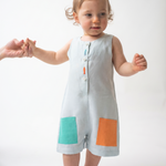 Bebê em pé usando Macaquinho Kalu de linho gelo, com bolsos coloridos e peixes bordados à mão.