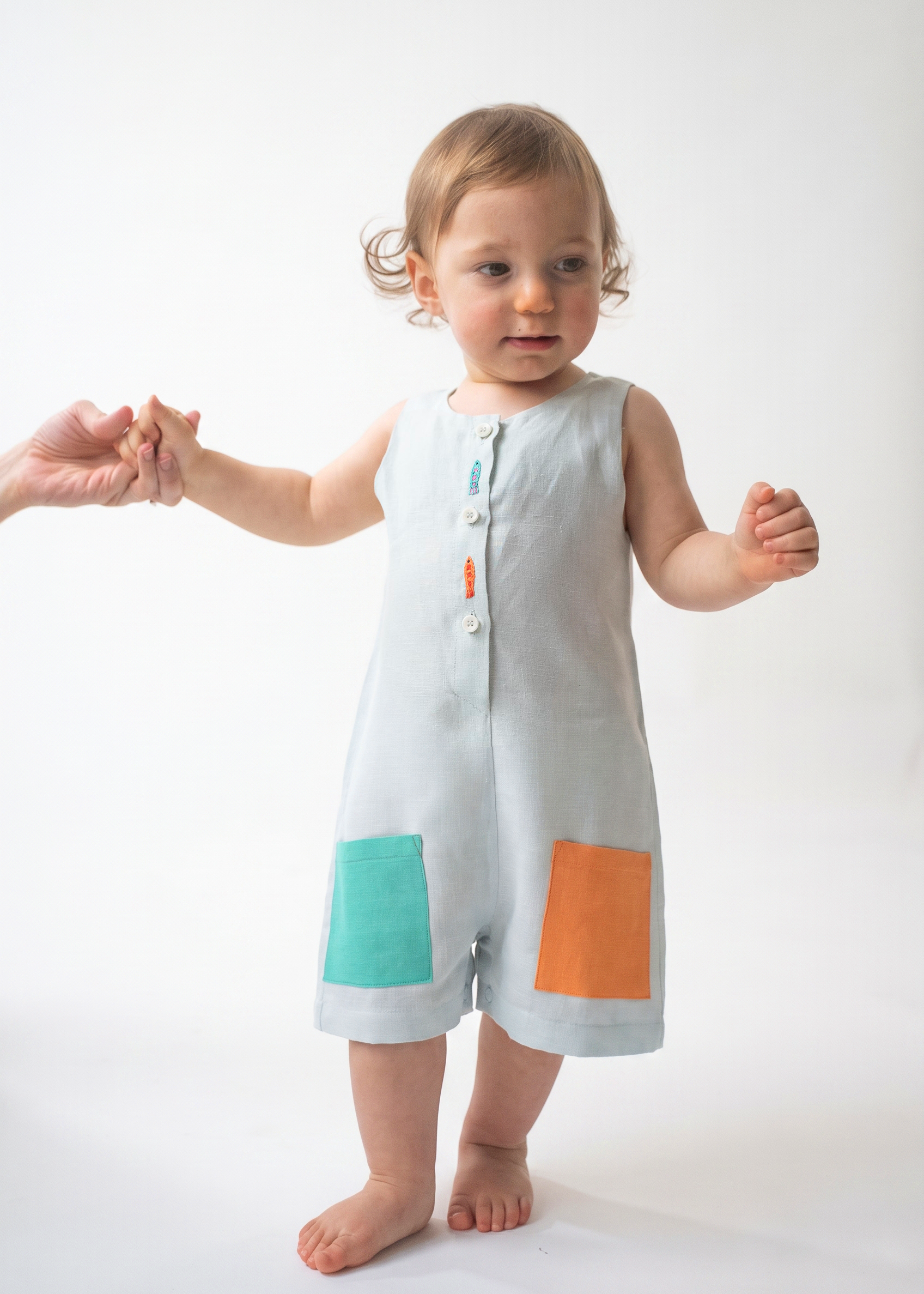 Bebê em pé usando Macaquinho Kalu de linho gelo, com bolsos coloridos e peixes bordados à mão.