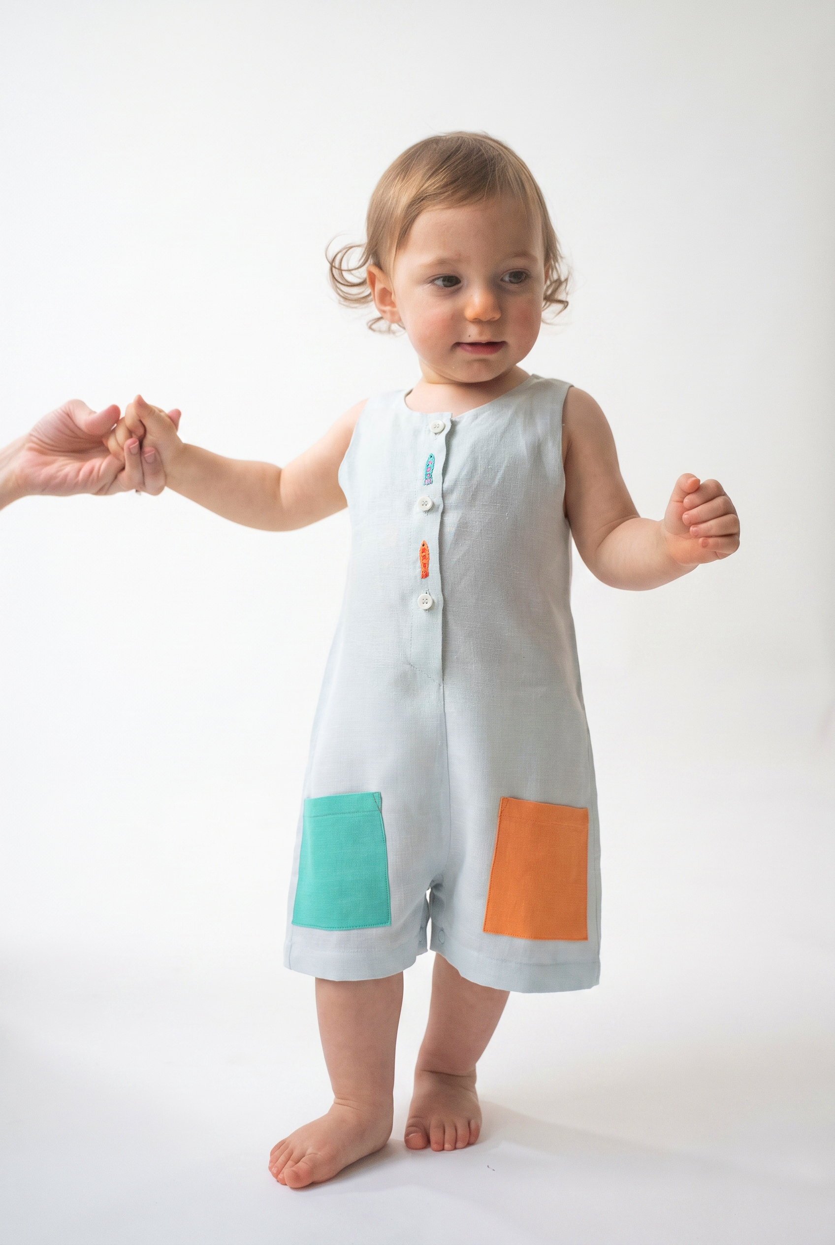 Bebê em pé usando Macaquinho Kalu de linho gelo, com bolsos coloridos e peixes bordados à mão.
