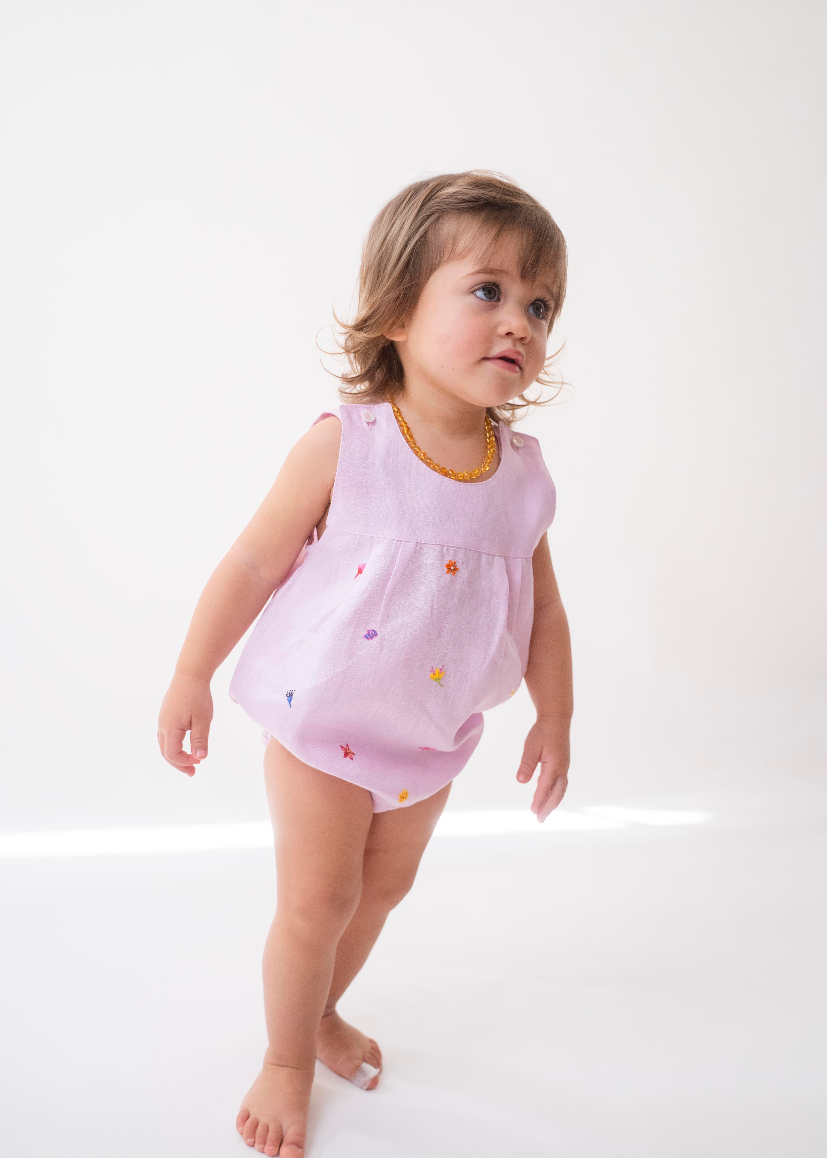 Bebê menina vestindo macaquinho infantil de linho rosa estilo romper, com flores coloridas bordadas à mão.