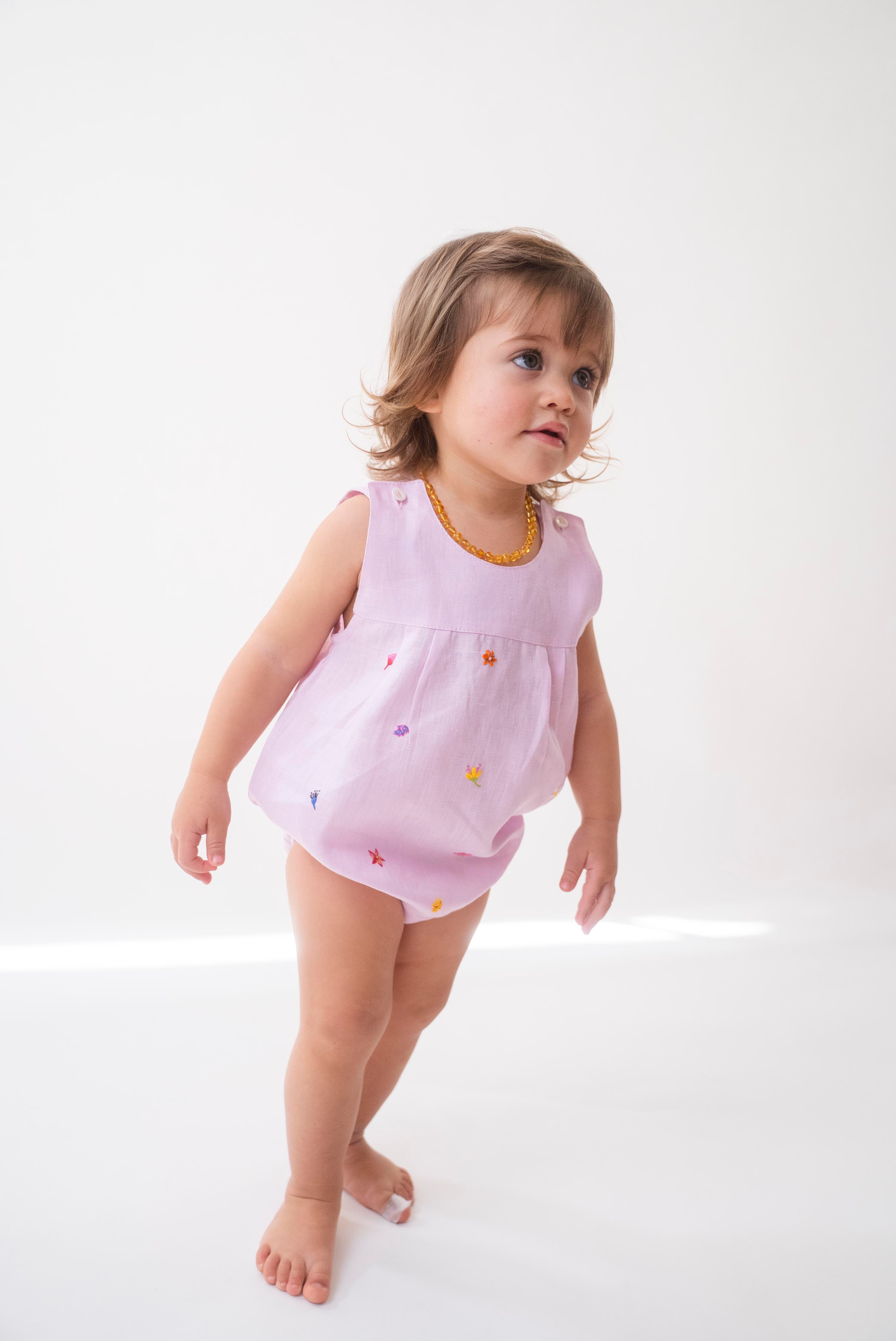 Bebê menina vestindo macaquinho infantil de linho rosa estilo romper, com flores coloridas bordadas à mão.