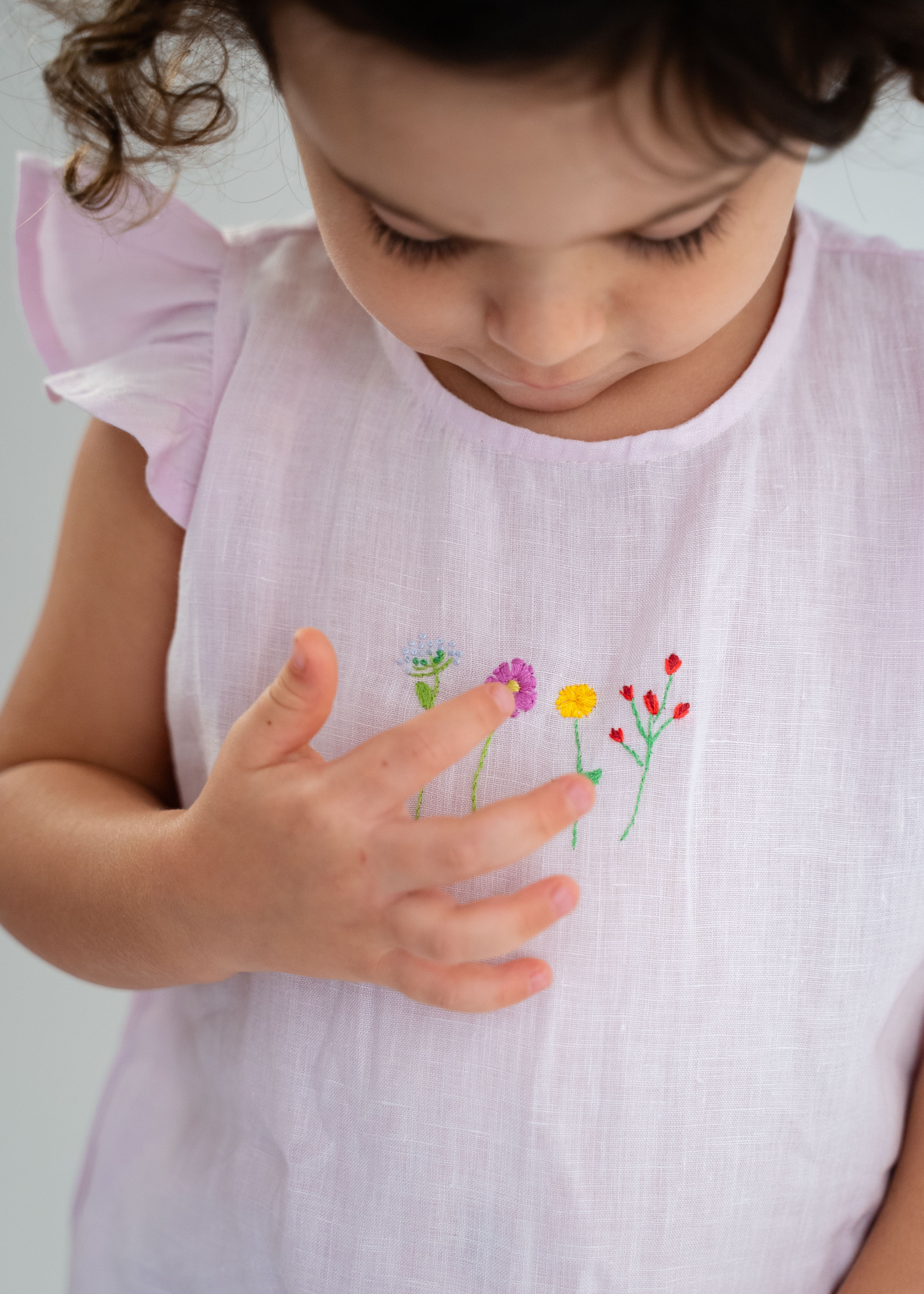 Bebê menina tocando com os dedos os bordado de flores do macaquinho estilo romper em linho cor de rosa.