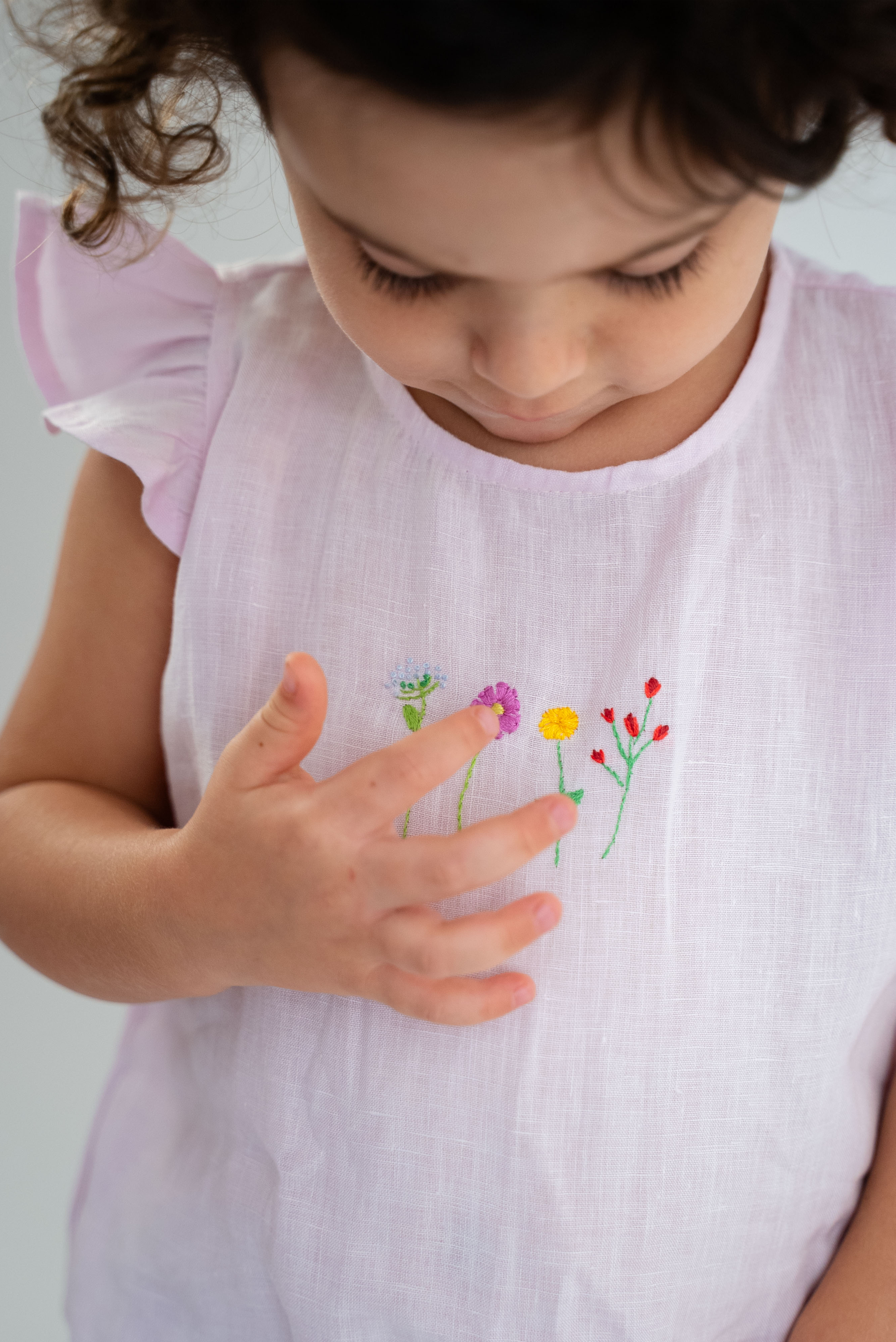 Bebê menina tocando com os dedos os bordado de flores do macaquinho estilo romper em linho cor de rosa.
