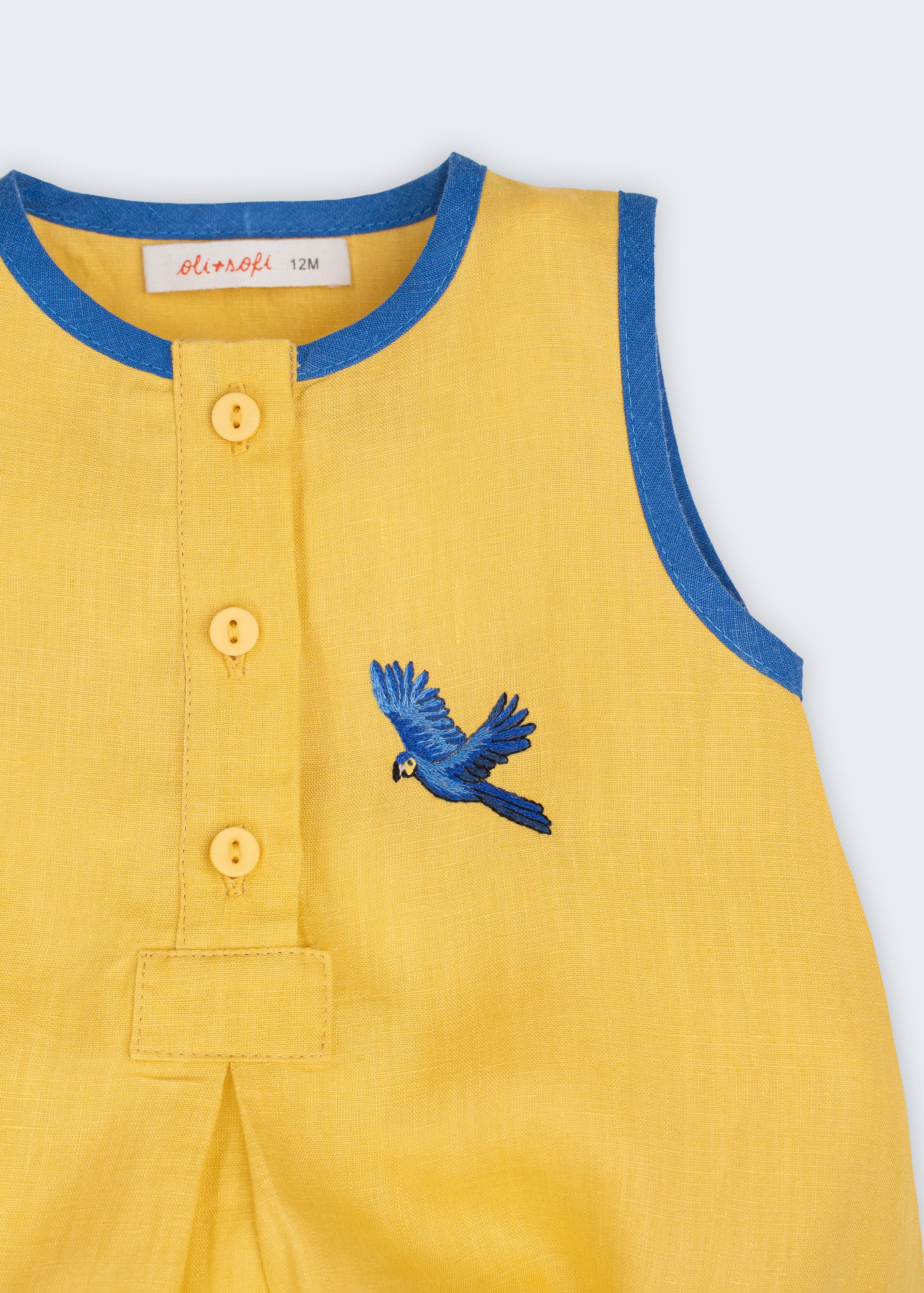 Macaquinho bebê em linho amarelo com acabamento azul e arara azul bordada à mão no peito, visto em detalhe